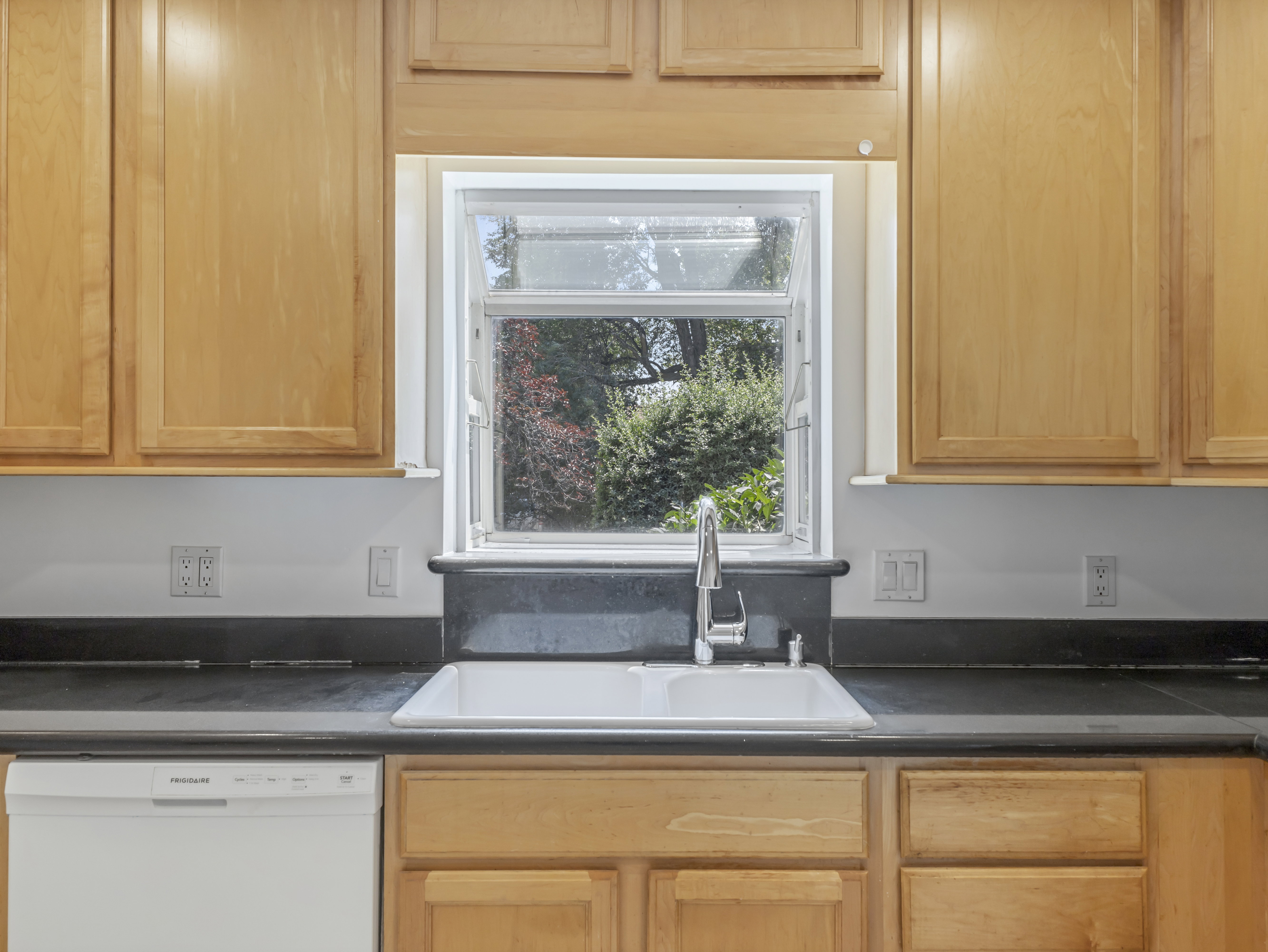 Kitchen sink area featuring light wood cabinetry, white wall paint, dark countertops, a white double basin sink, and a stainless steel faucet