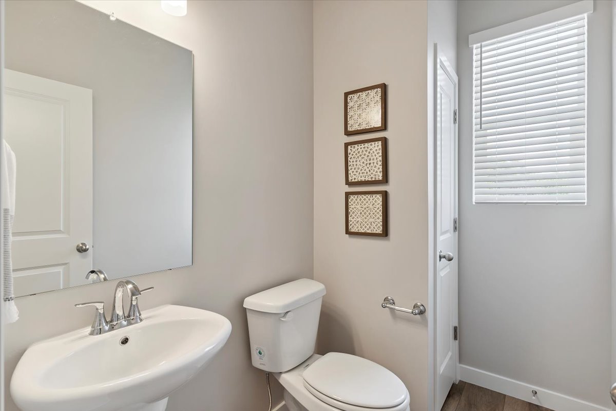 Pedestal sink with chrome faucet, a rectangular mirror, a white toilet, and wood-finish flooring