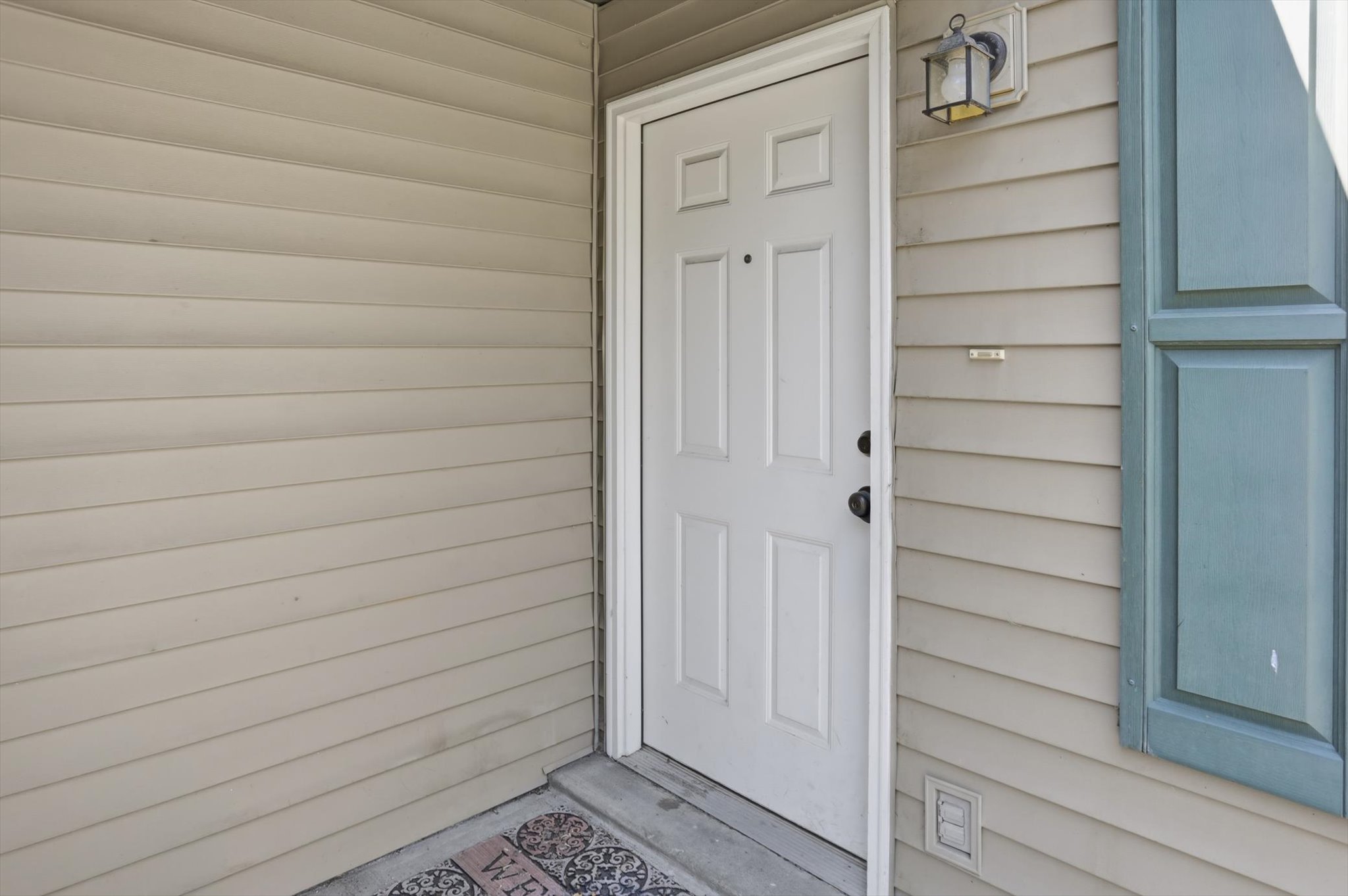 Entry with a four-panel door, tan horizontal siding, and a mounted exterior light fixture