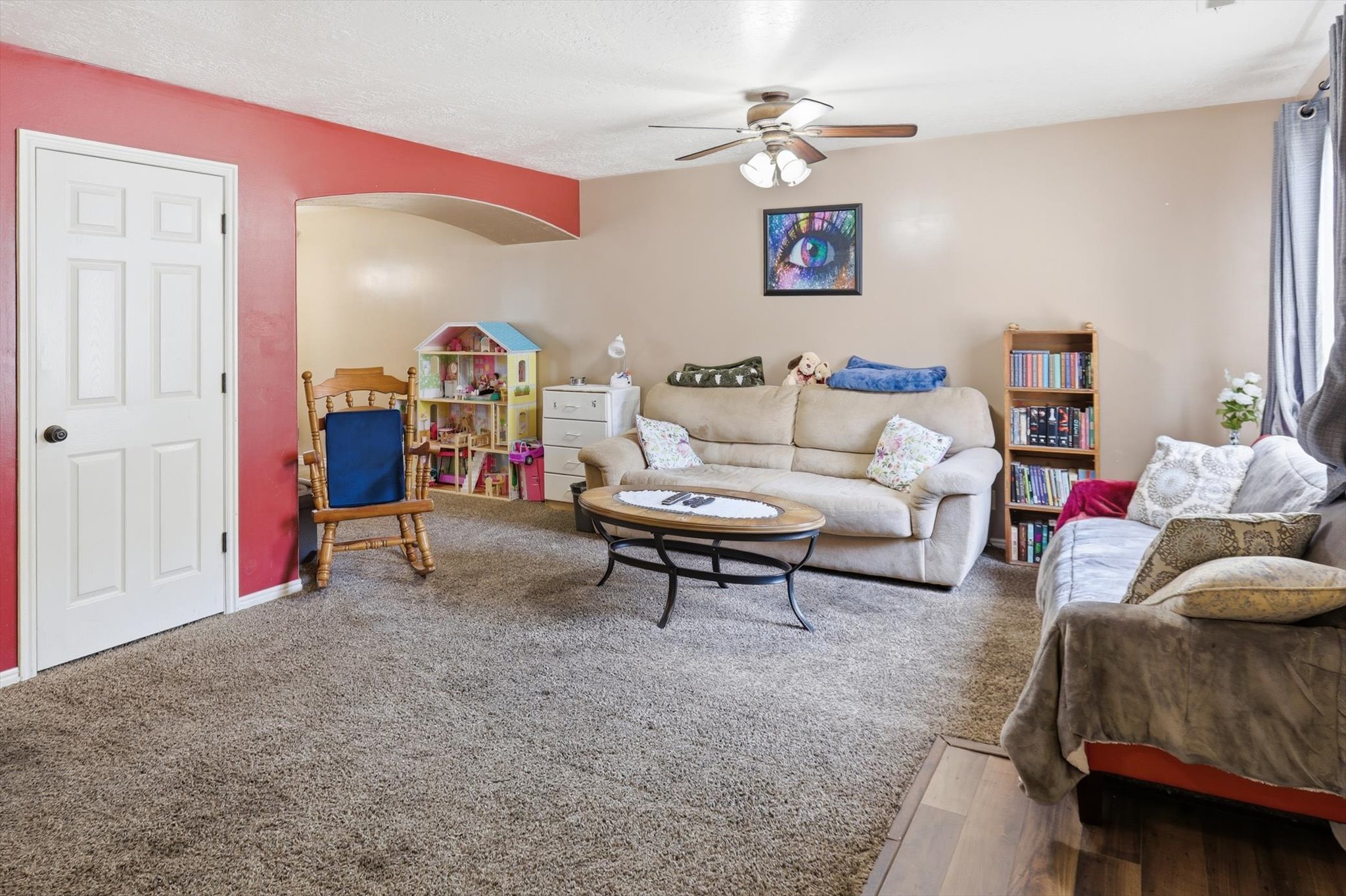 Carpeted living area featuring a ceiling fan with integrated lighting, an arched doorway, a white paneled door, and a defined section of wood-finish flooring