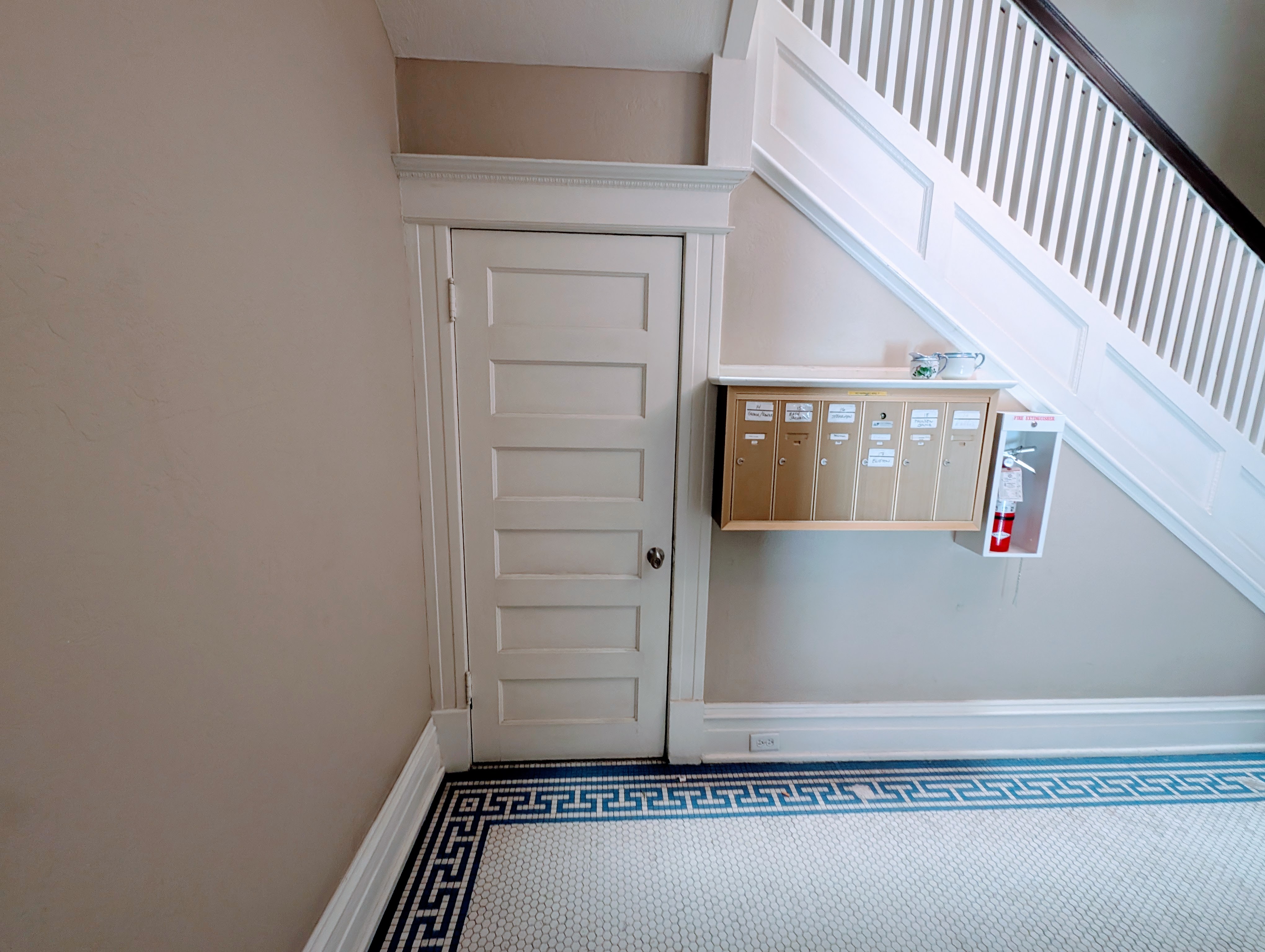Entryway featuring classic paneled door with detailed trim, a built-in mail station, staircase with white balusters and dark handrail, and mosaic tile flooring with a Greek key border
