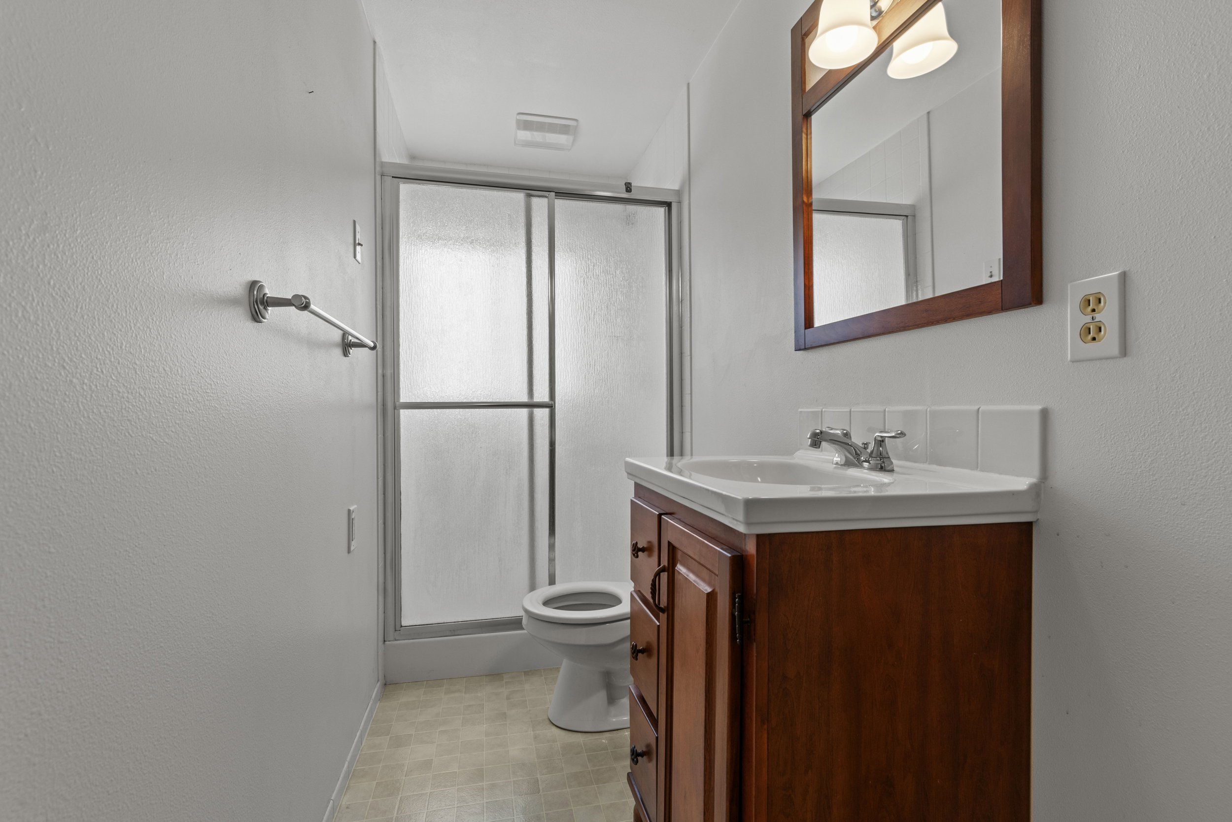 Bathroom featuring a wood-finish vanity with an integrated sink, a rectangular framed mirror with sconce lighting, a shower with sliding frosted glass doors, and light-toned vinyl flooring