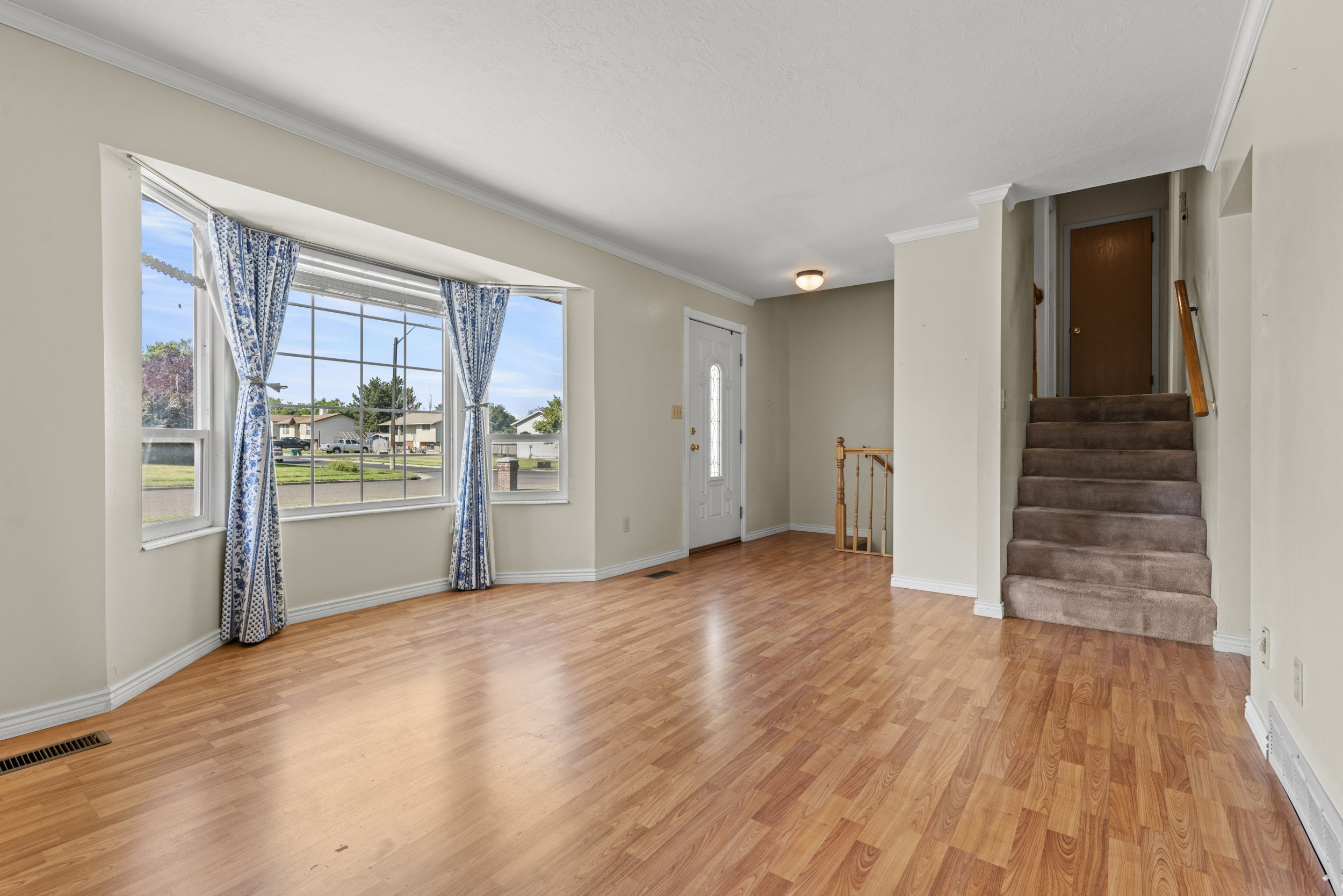 Spacious interior featuring wood-finish flooring, a prominent bay window, neutral wall tones, white trim, and a carpeted staircase