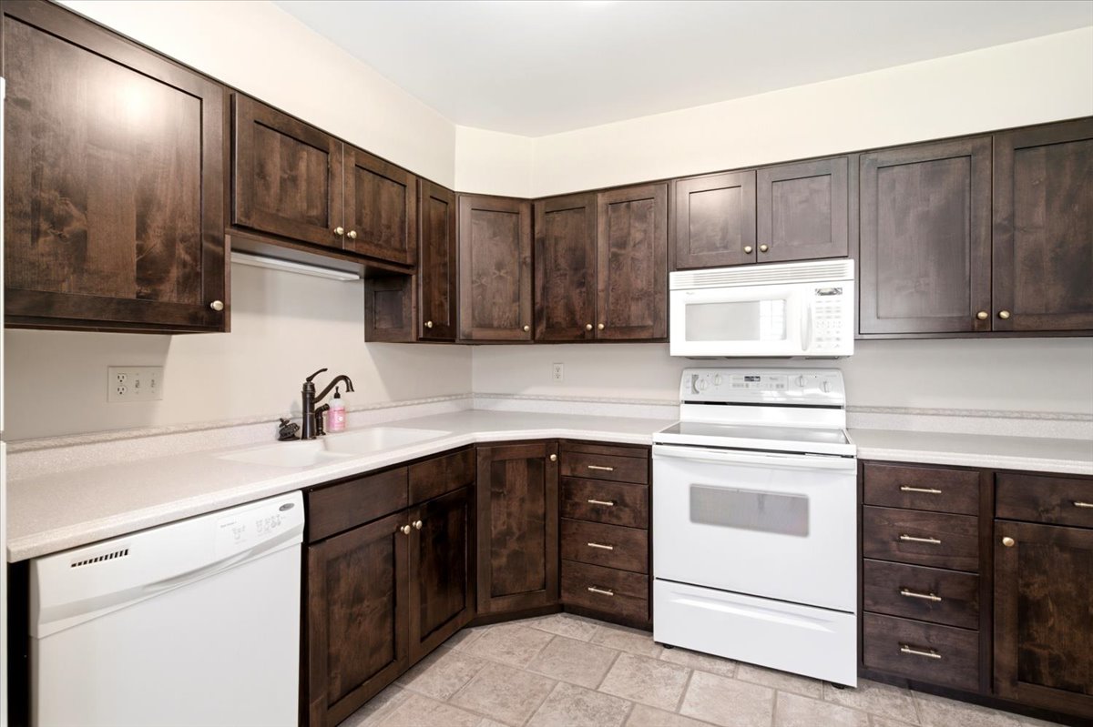 Kitchen featuring dark wood-finish cabinetry, light-toned countertops, white appliances, a double basin sink, and tile flooring