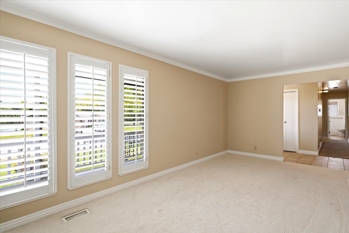 Spacious interior room featuring three windows with white plantation shutters, crown molding, and neutral-toned carpeting