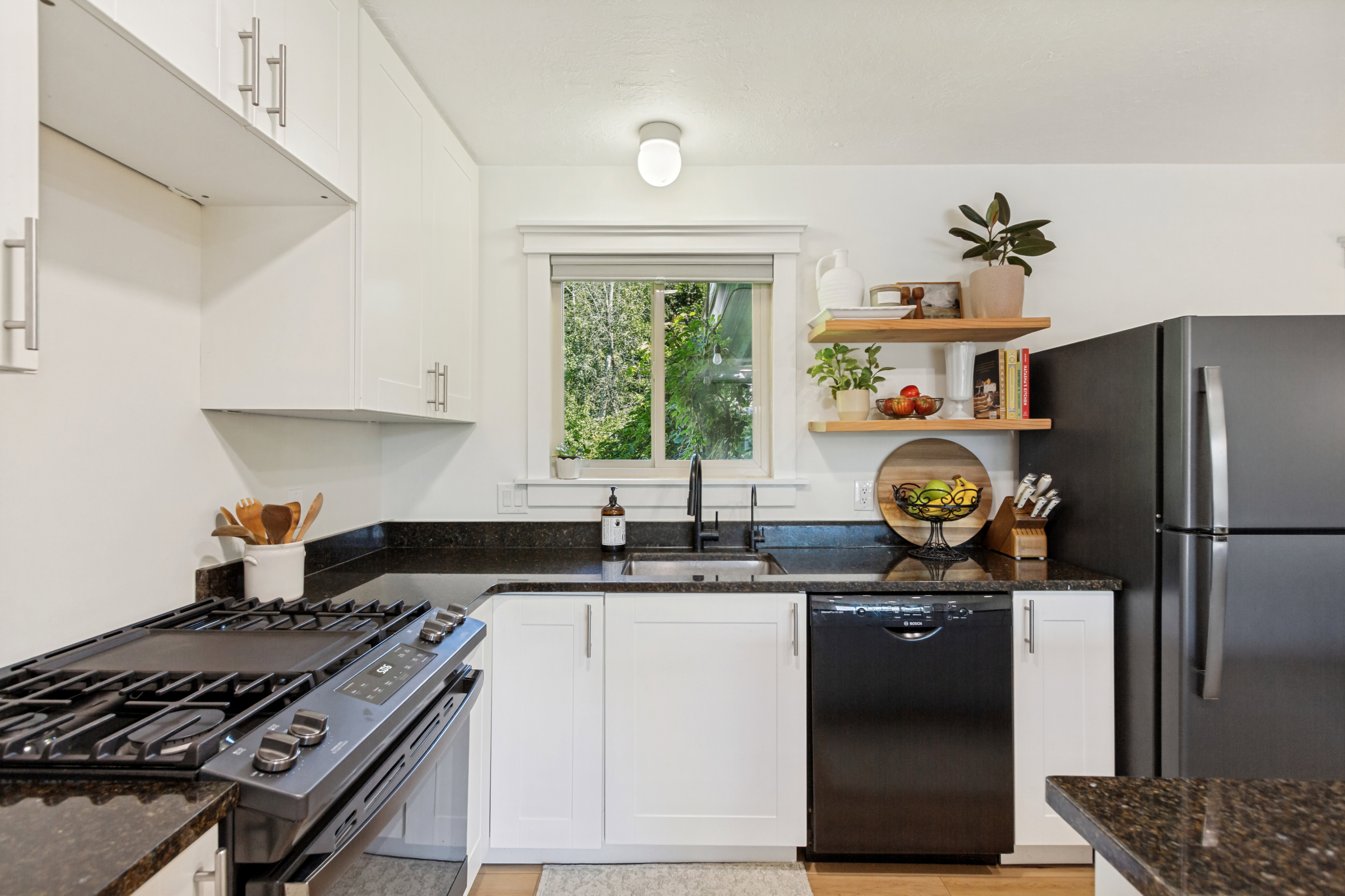 Well-appointed kitchen featuring white cabinetry, dark granite countertops, stainless steel sink with a black faucet, black appliances, and open wood shelving