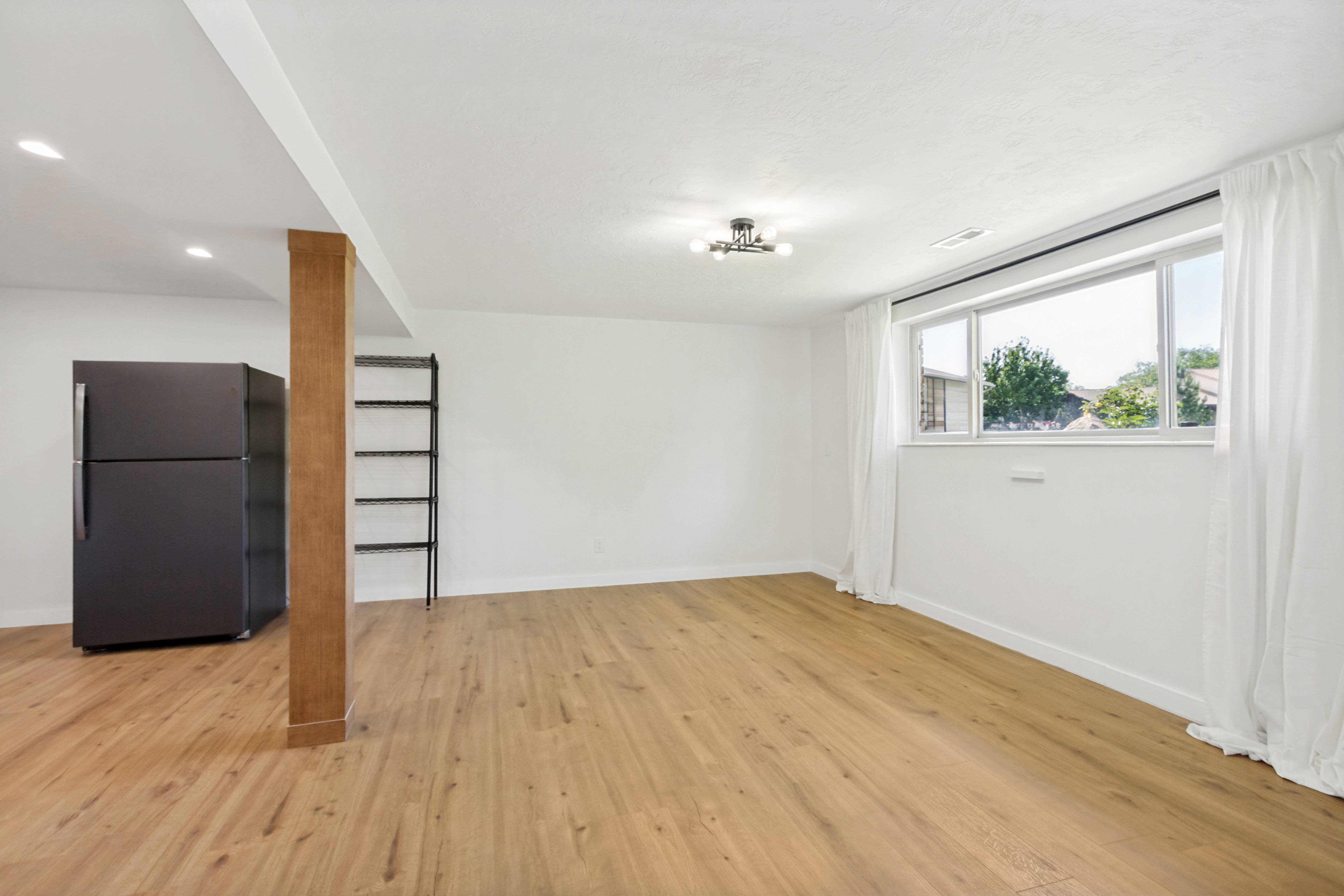 Spacious room featuring wood-finish flooring, a large window with white curtains, and recessed lighting
