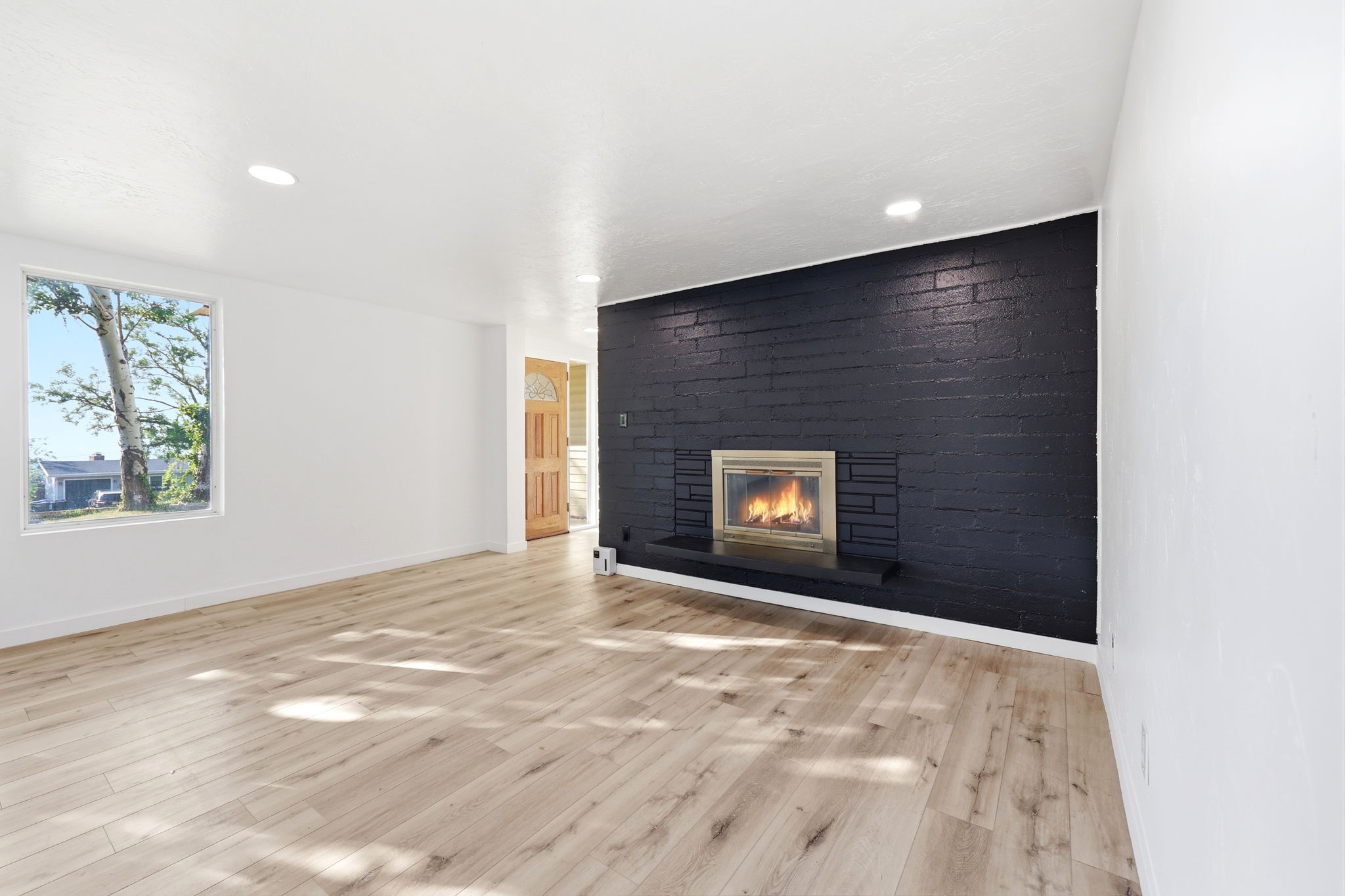 Living room featuring a brick fireplace with a dark finish, a light wood-finish floor, a rectangular window, recessed lighting, and a wood entry door with glass inserts