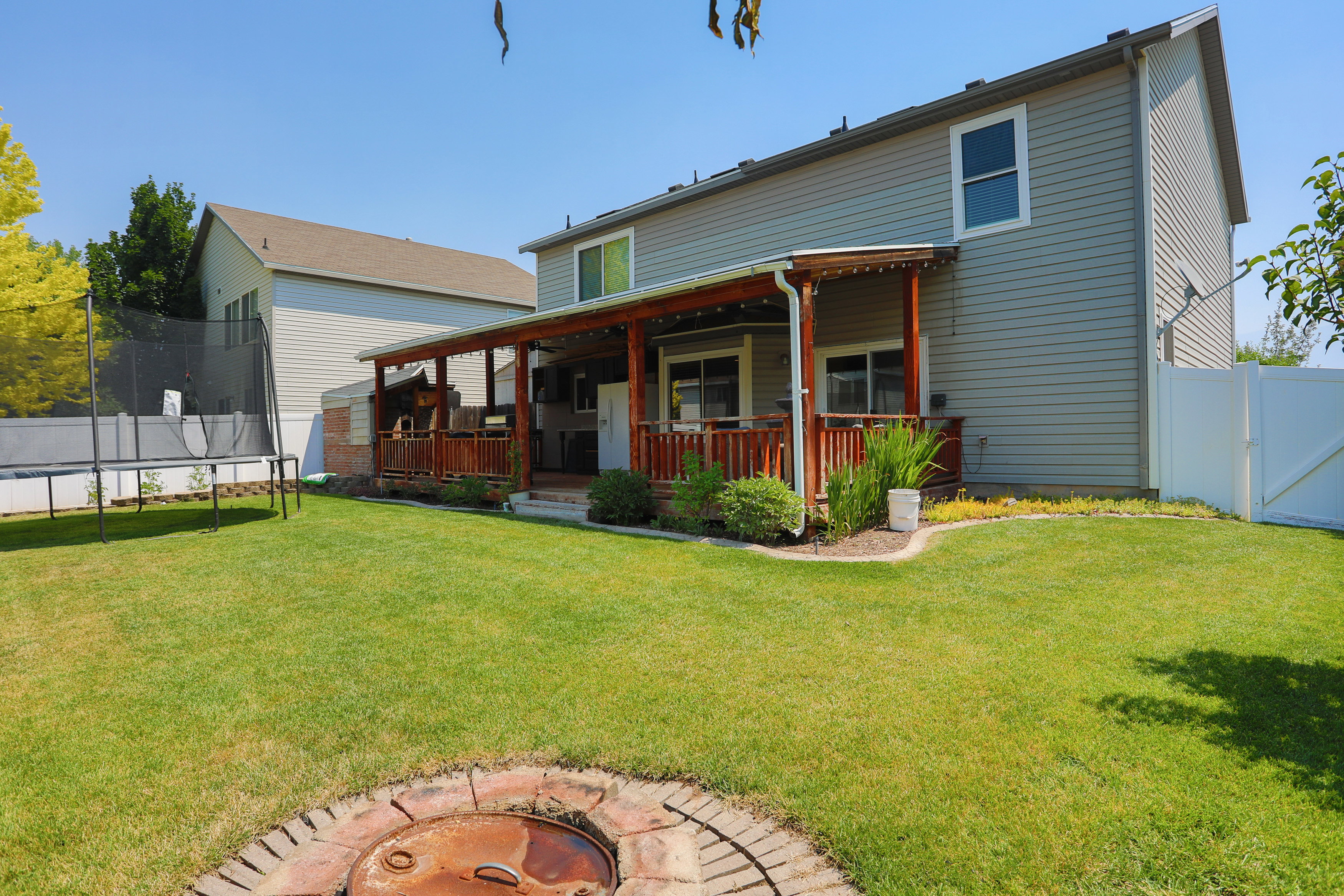 Spacious backyard featuring a covered patio, expansive lawn, mature landscaping, and a paver-lined fire pit