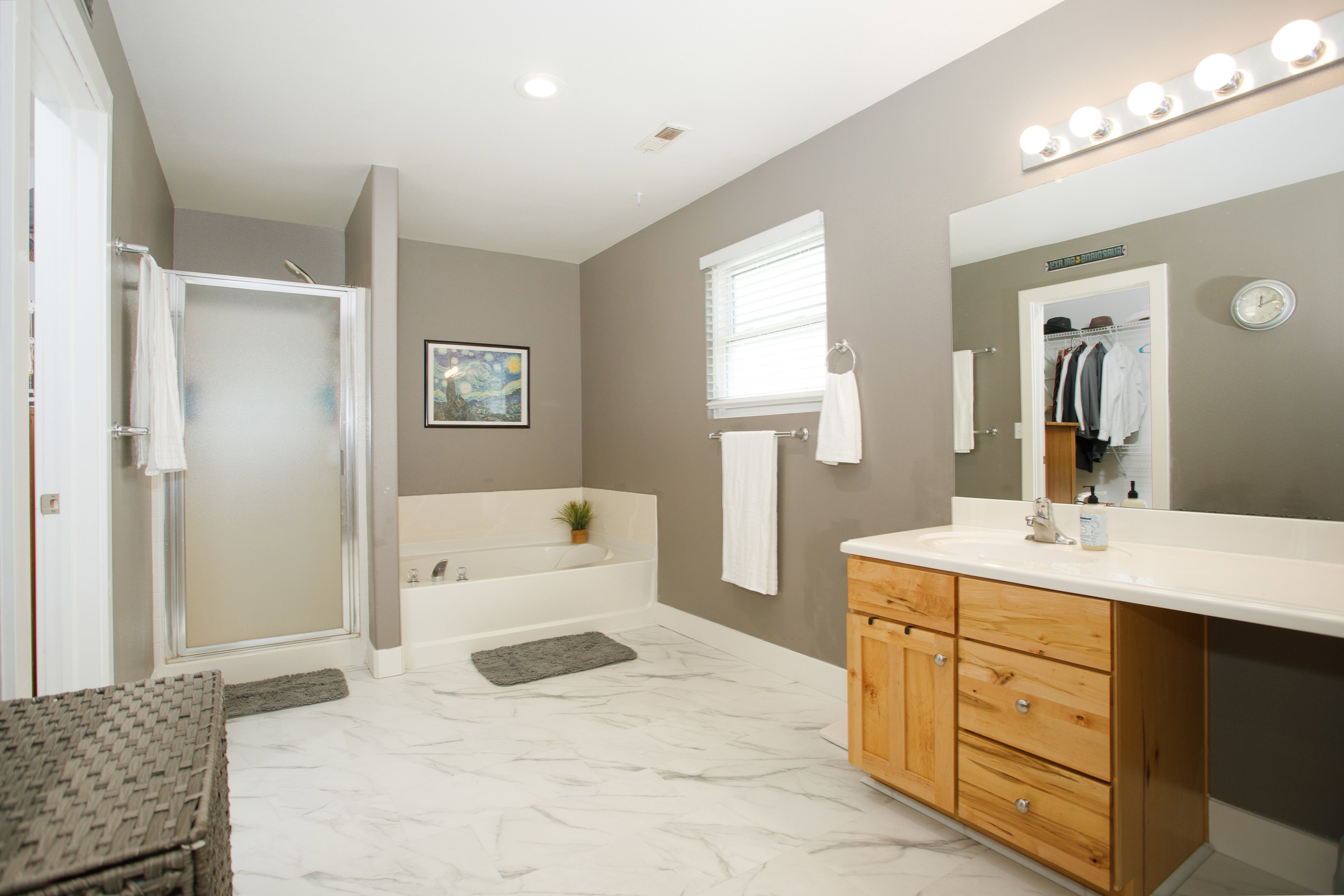 Spacious bathroom featuring large format tile flooring, a glass-enclosed shower, a built-in soaking tub, a single vanity with a light wood-finish cabinet, and a large mirror