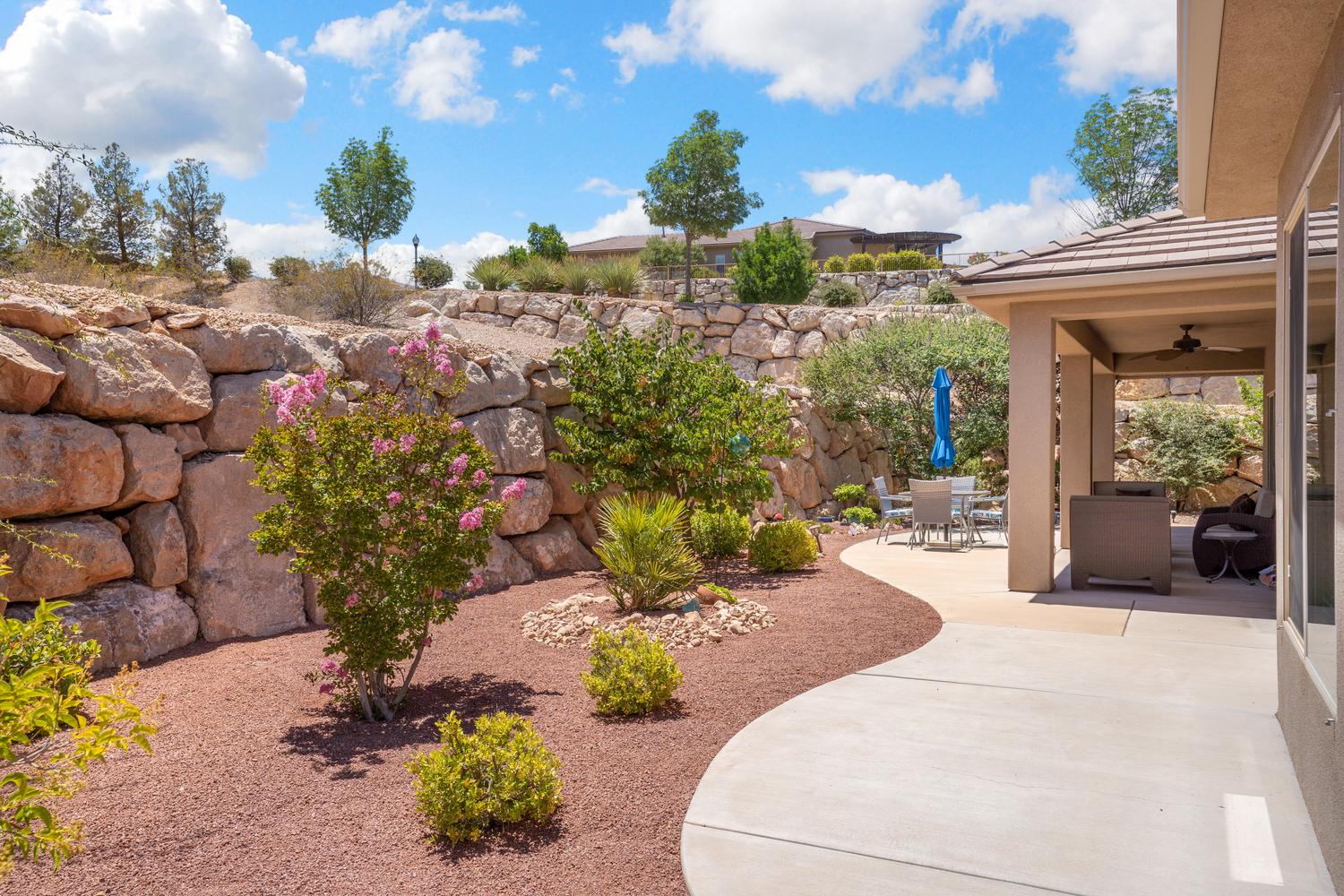Expansive outdoor living area featuring a covered patio with ceiling fan, curved concrete pathways, and xeriscaping with mature plantings