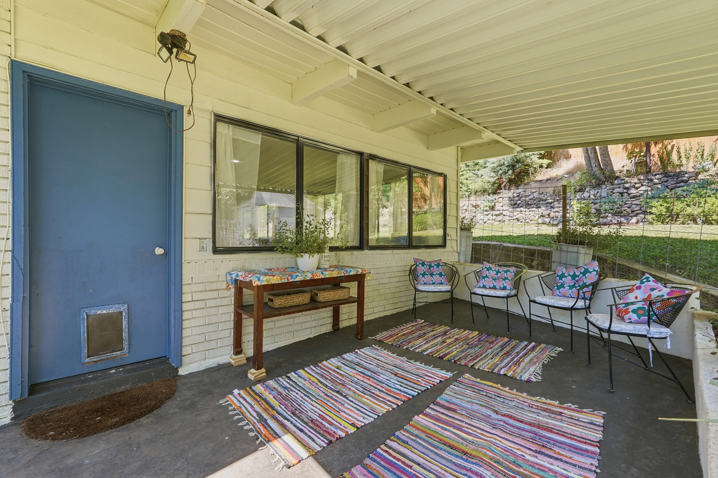 Covered outdoor patio featuring a solid blue door, white brick siding, two large windows, and a sloped, treed backdrop