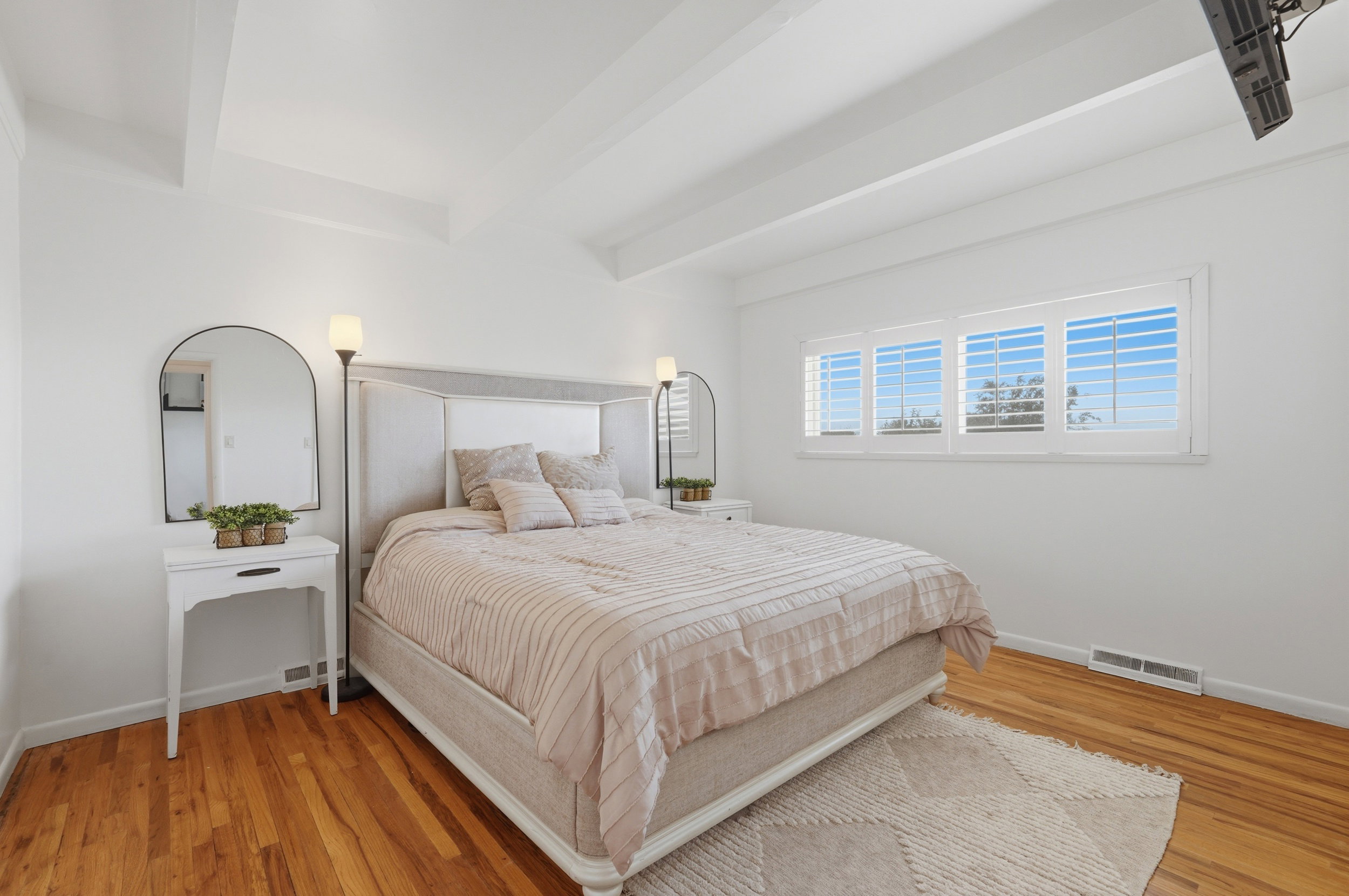 Spacious primary bedroom featuring warm wood-finish flooring, a tray ceiling with exposed beams, and a wide window with plantation shutters