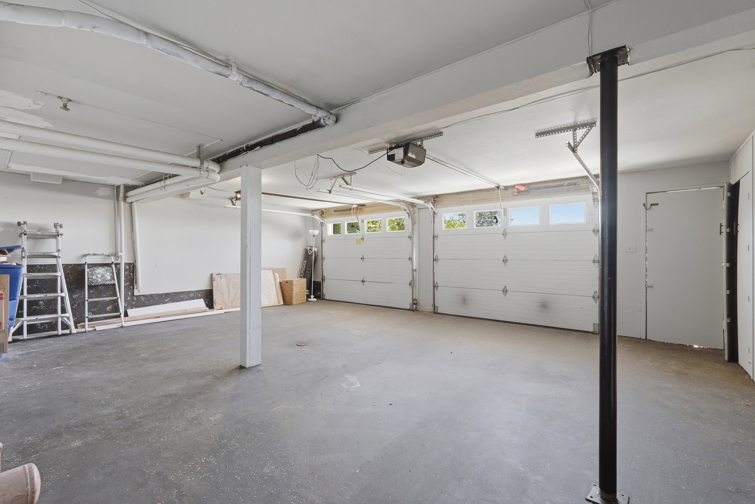 Spacious garage featuring two overhead garage doors with window panels, concrete flooring, exposed ceiling beams, and a side door for additional access