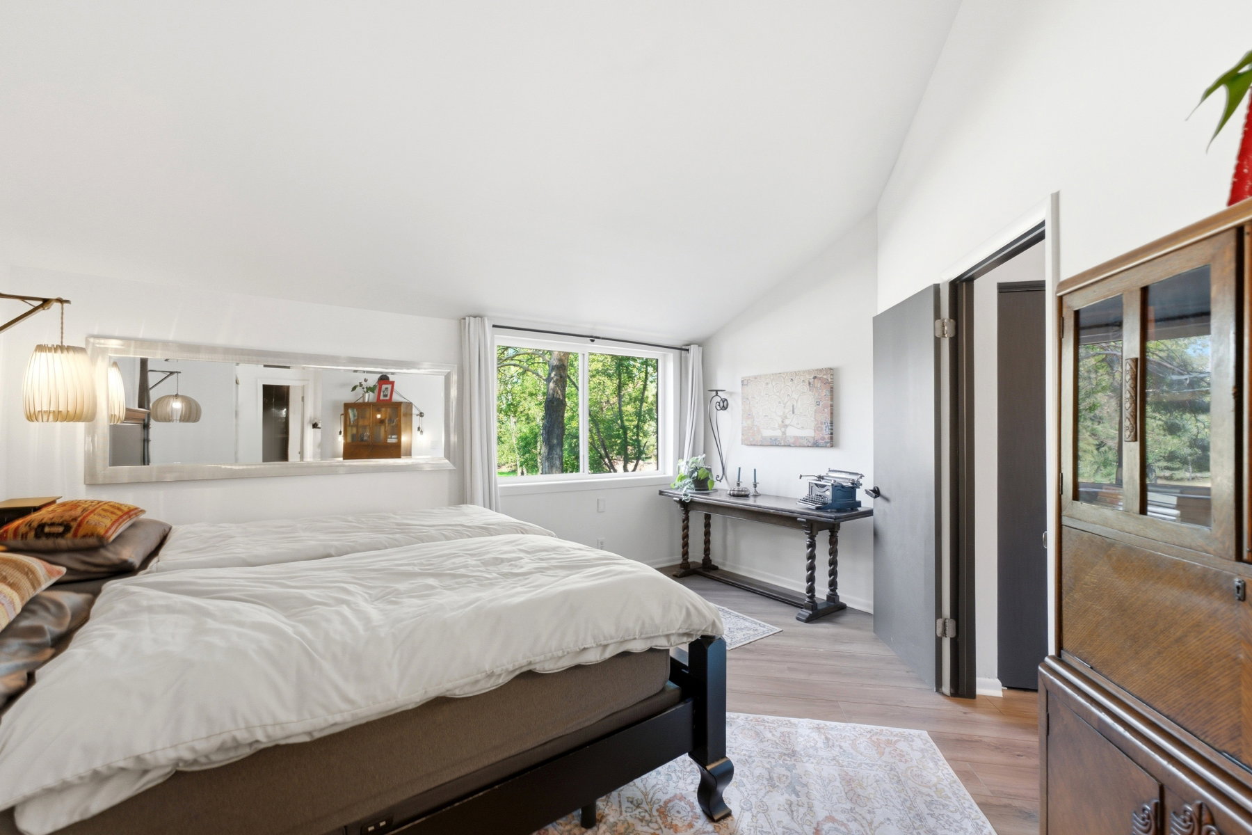 Vaulted ceiling bedroom with wood-finish flooring, a large window, a built-in mirror, and a modern wall-mounted light fixture
