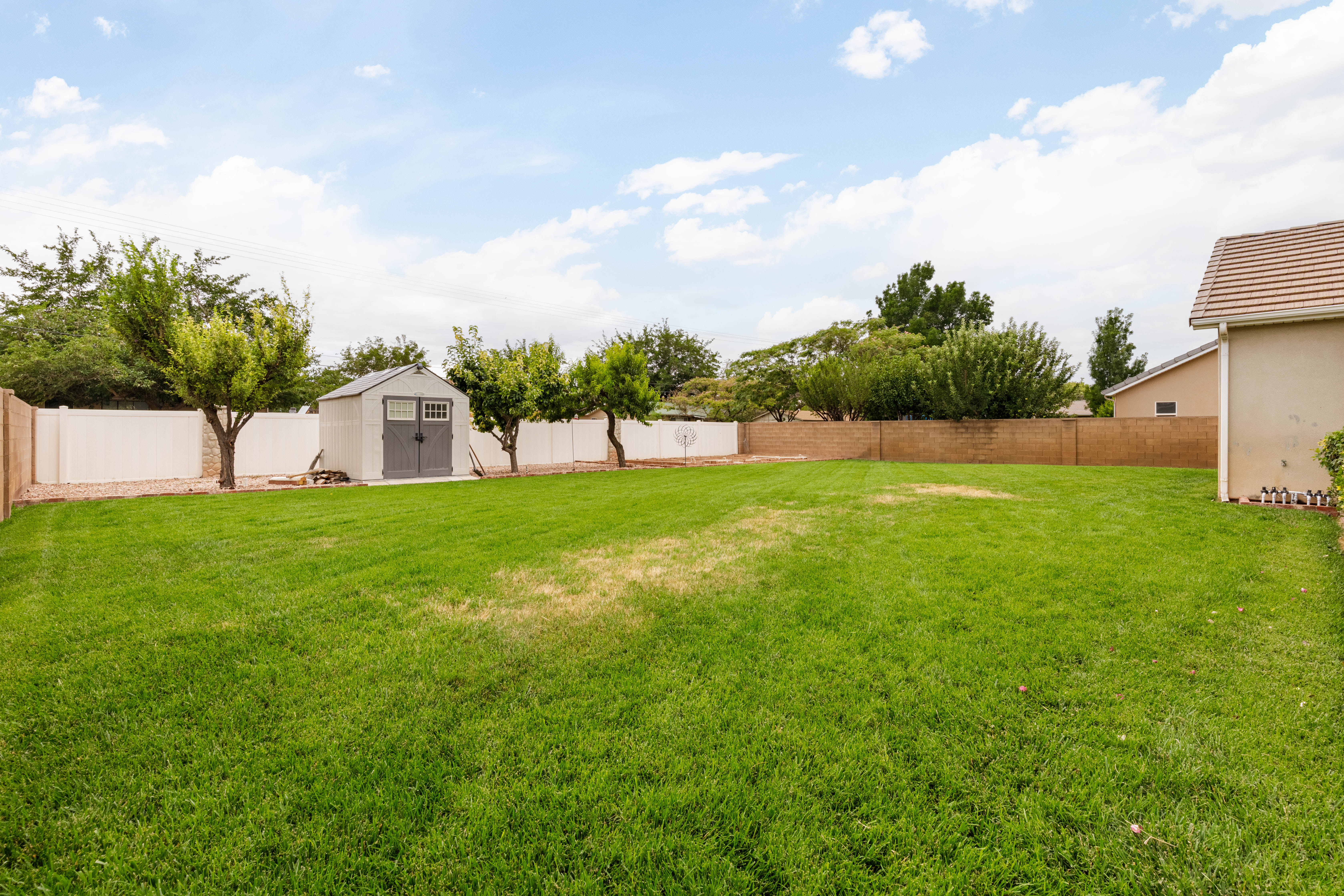 Expansive grass backyard featuring a detached storage shed, mature trees, and a combination of white vinyl and brick fencing