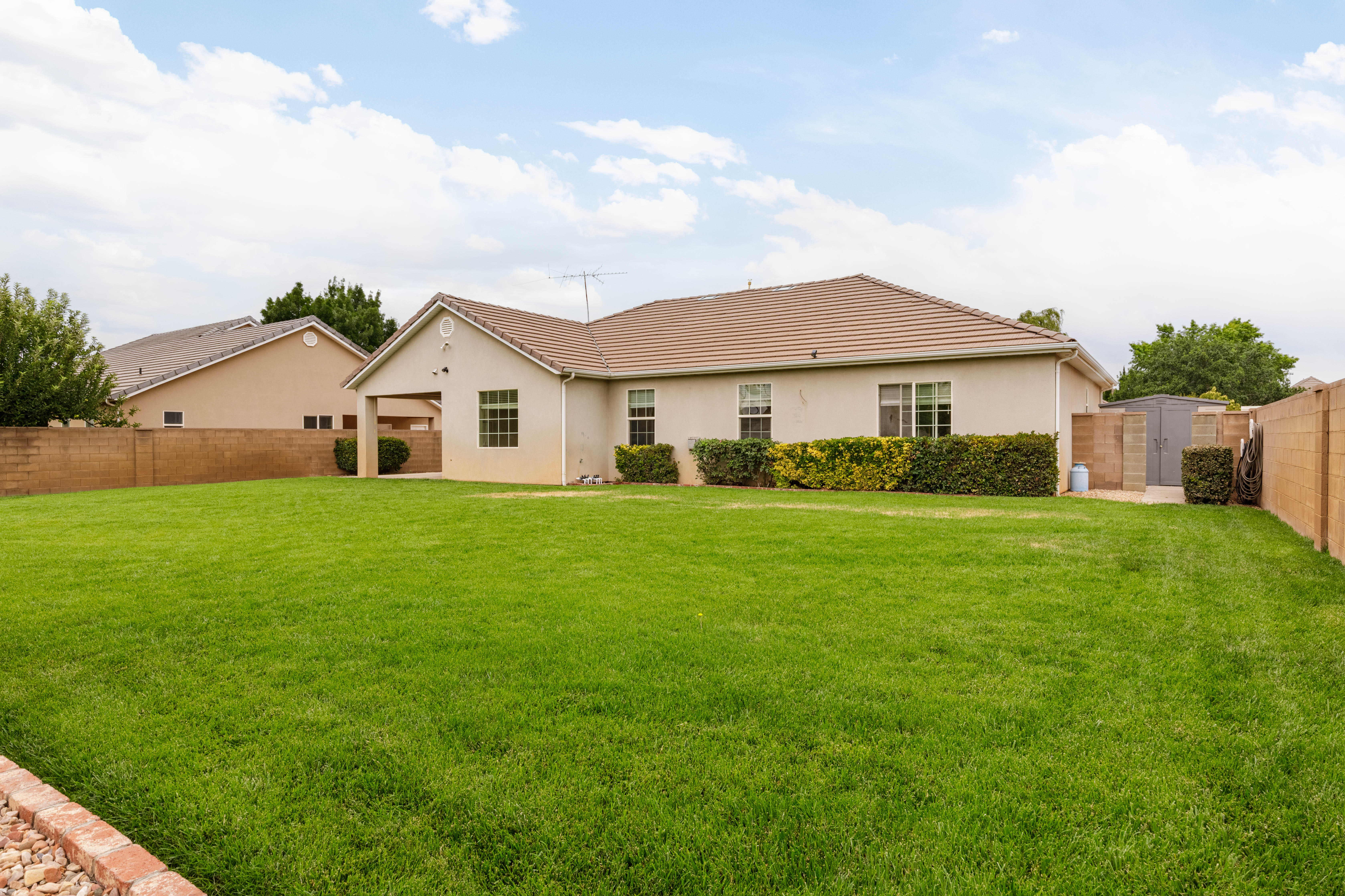 Expansive backyard featuring a large green lawn, concrete patio, stucco exterior, tan tiled roofing, and a storage shed