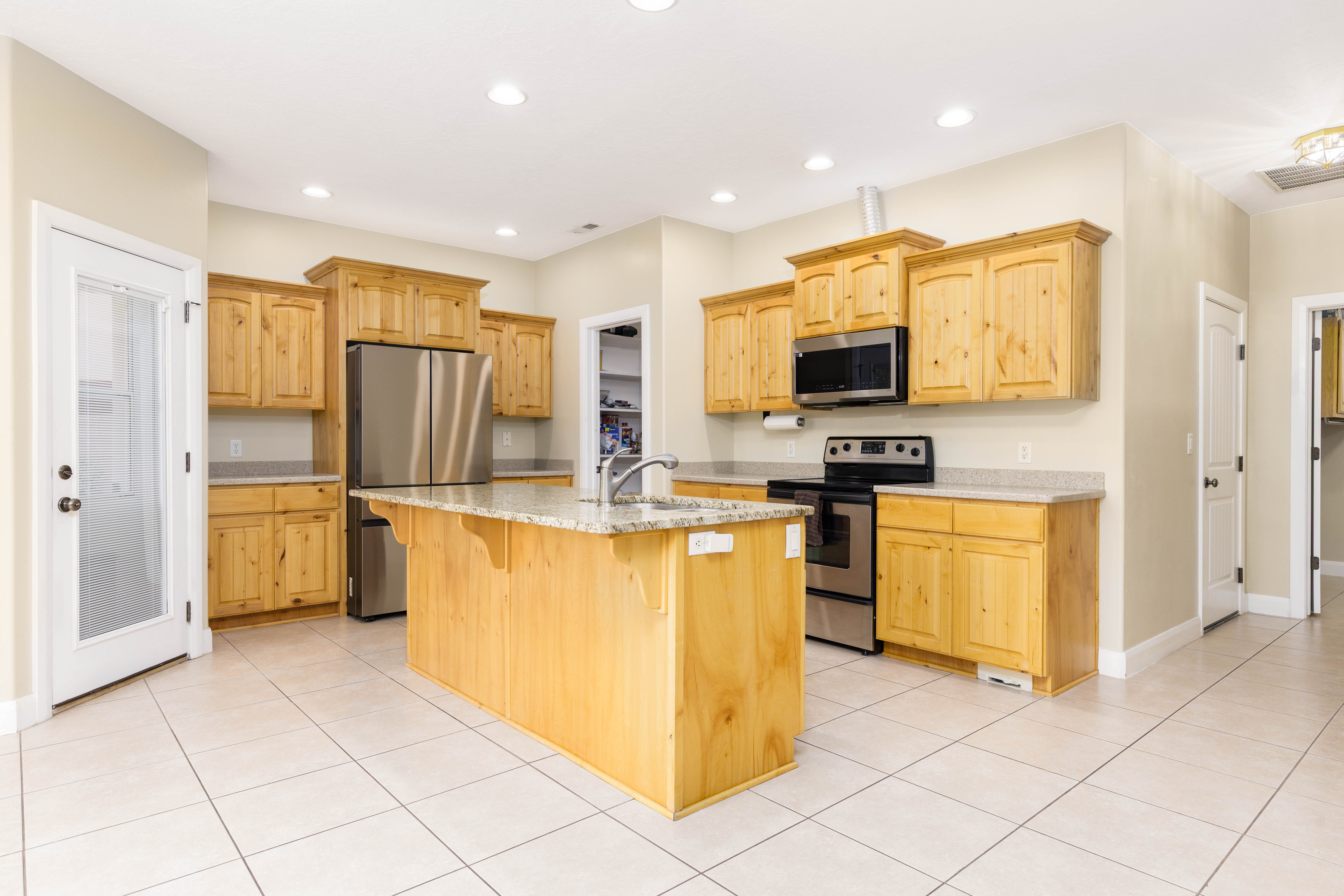 Spacious kitchen featuring light wood cabinetry, a central island with a light-colored countertop, stainless steel appliances, and large format tile flooring