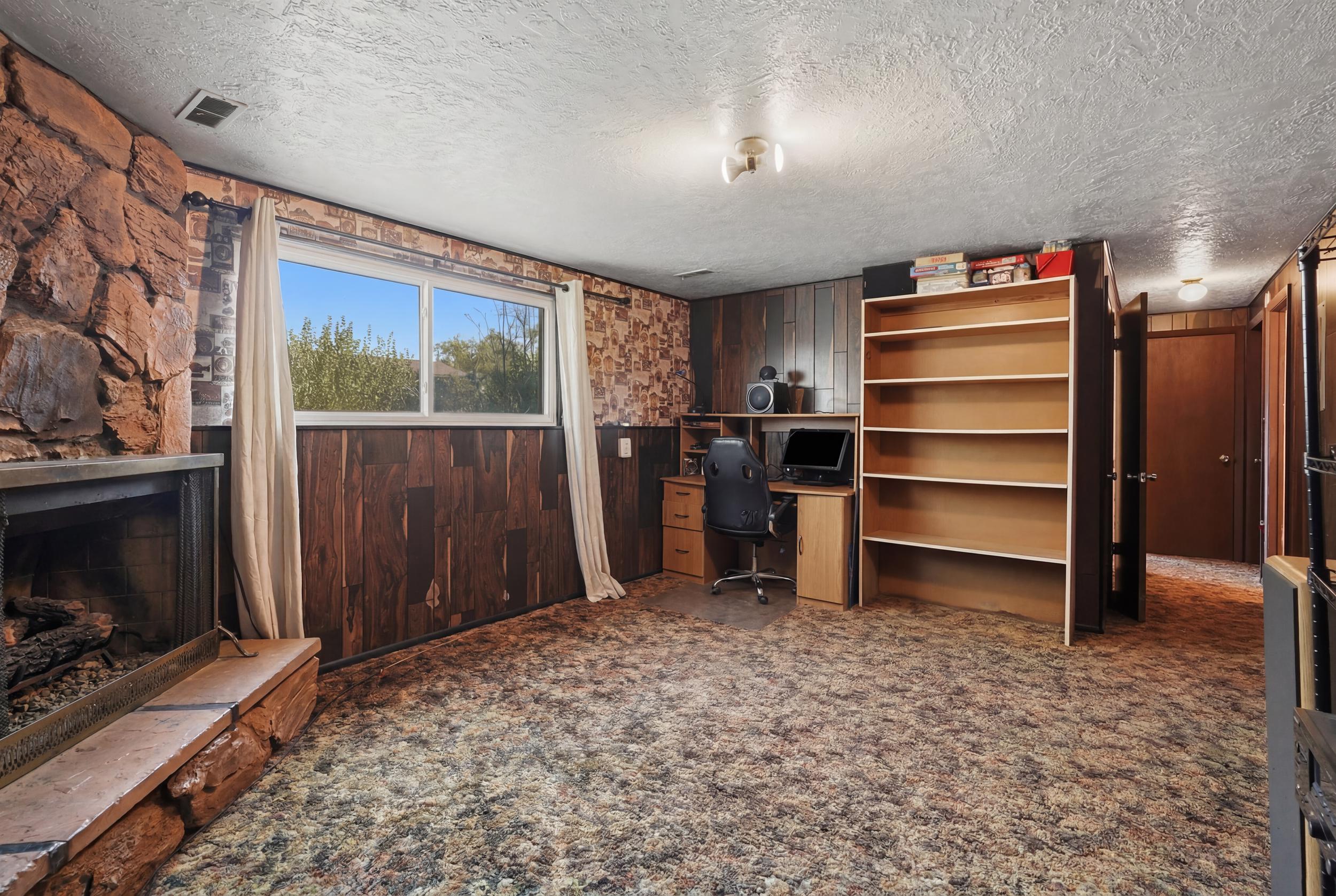 Spacious room featuring a stone fireplace with hearth, a desk area with built-in cabinetry, an expansive window, and a large built-in shelving unit