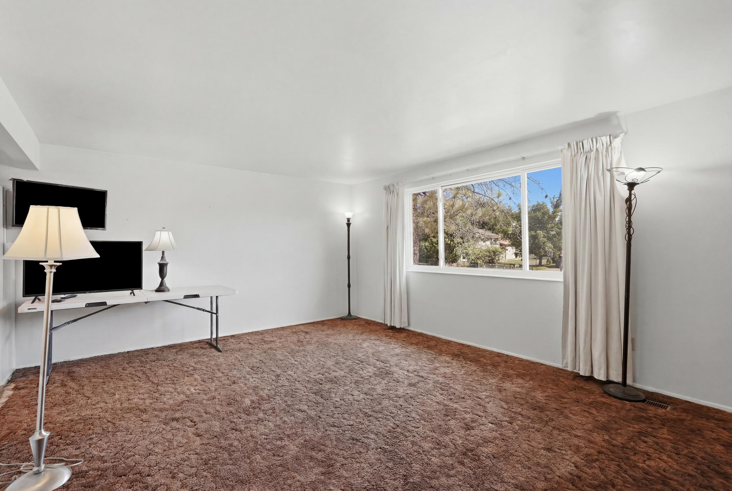 Spacious room featuring broadloom carpeting, crisp white walls, and a large window with white trim and drapery