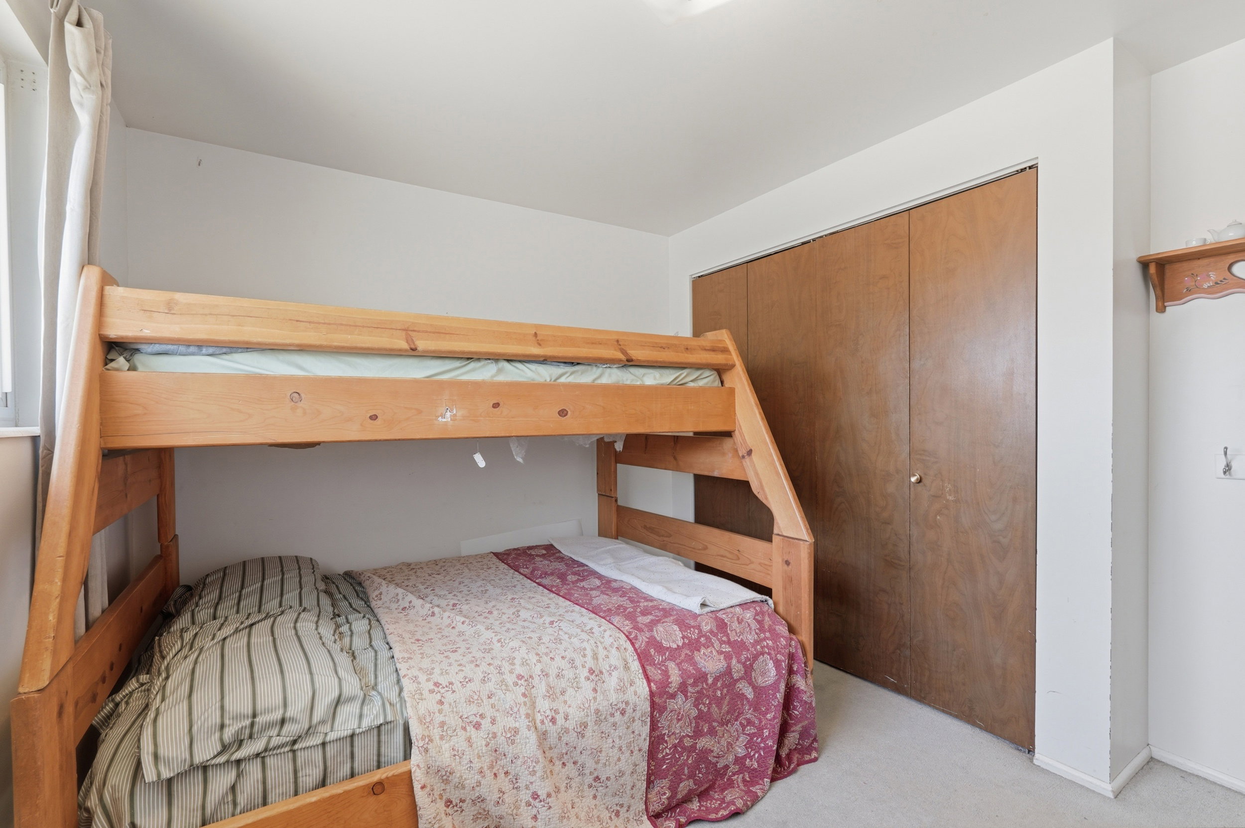 Carpeted room with a wooden bunk bed frame, wood-finish bi-fold closet doors, and a window with white drapes