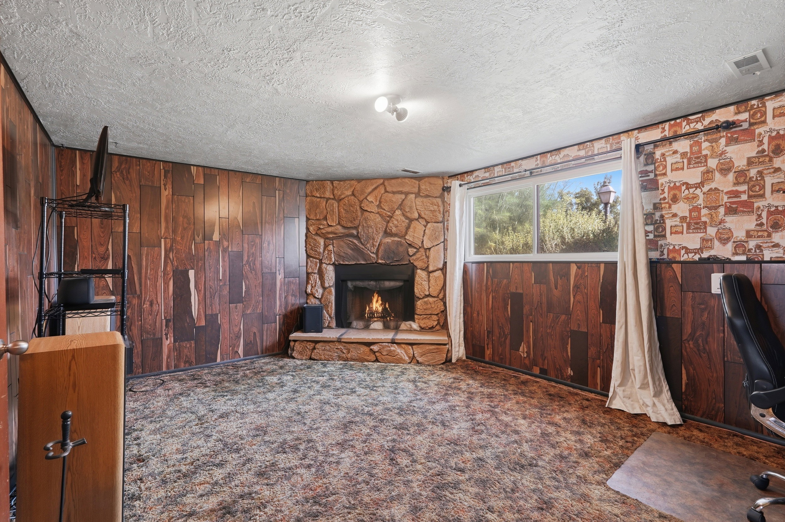 Corner stone fireplace with hearth, wood-panel accent walls, and a large window providing natural light