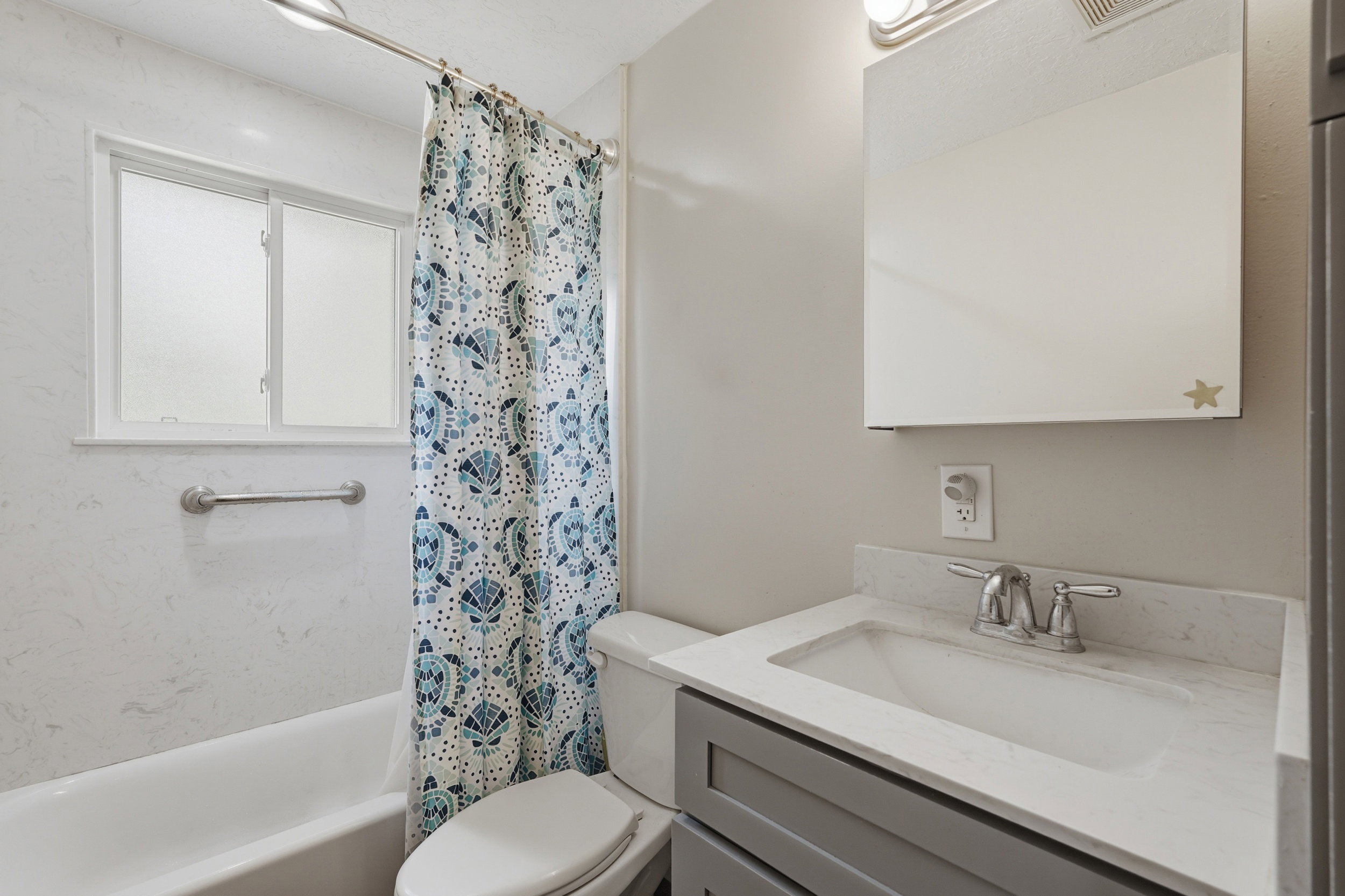 Contemporary bathroom featuring a white bathtub, frosted window, chrome grab bar, vanity with gray cabinetry, and a white countertop with an integrated sink