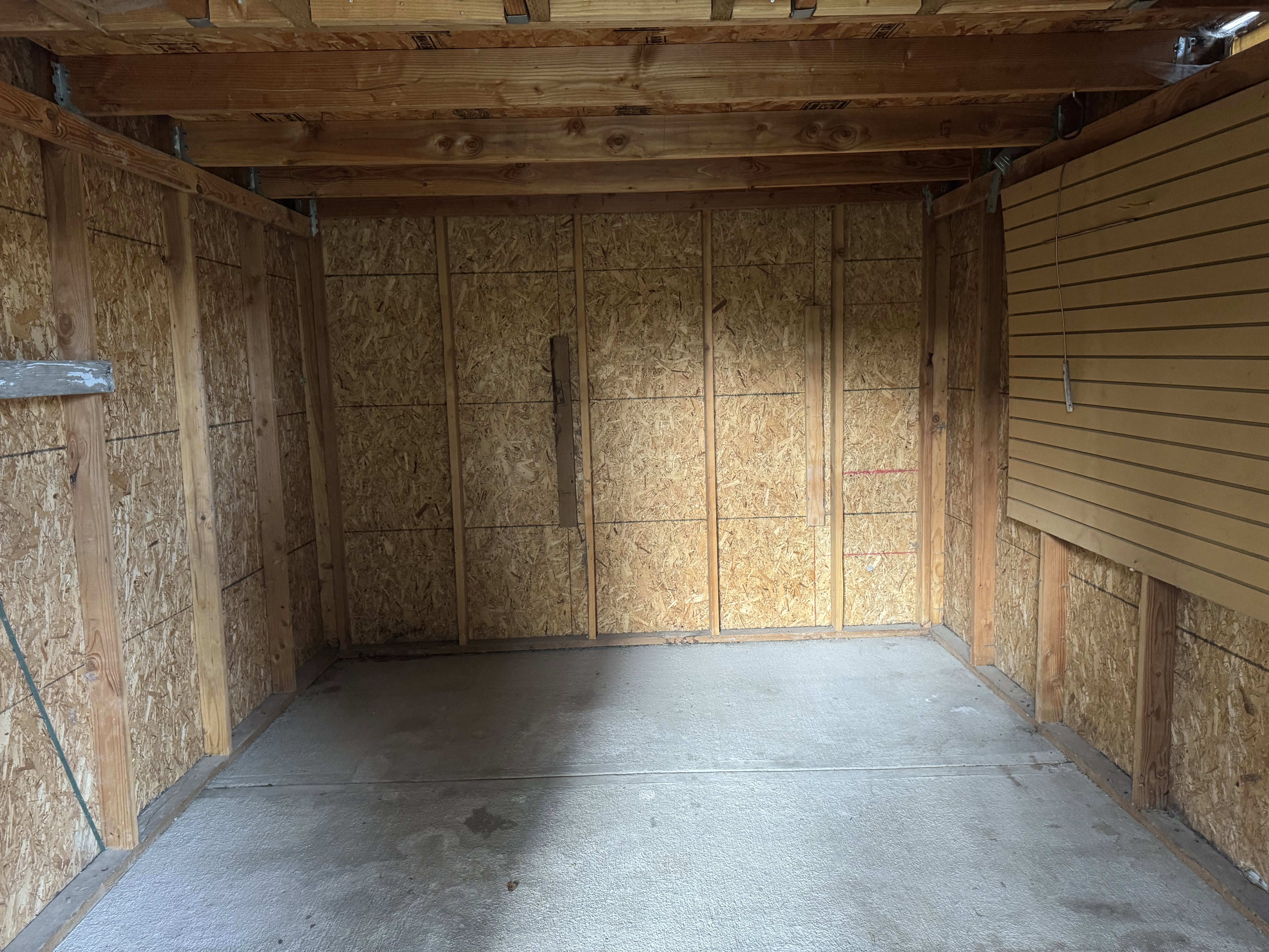 Storage shed is 10 feet by 12 feet. featuring a concrete floor, exposed wooden ceiling beams, oriented strand board walls, and a slatwall panel for organization