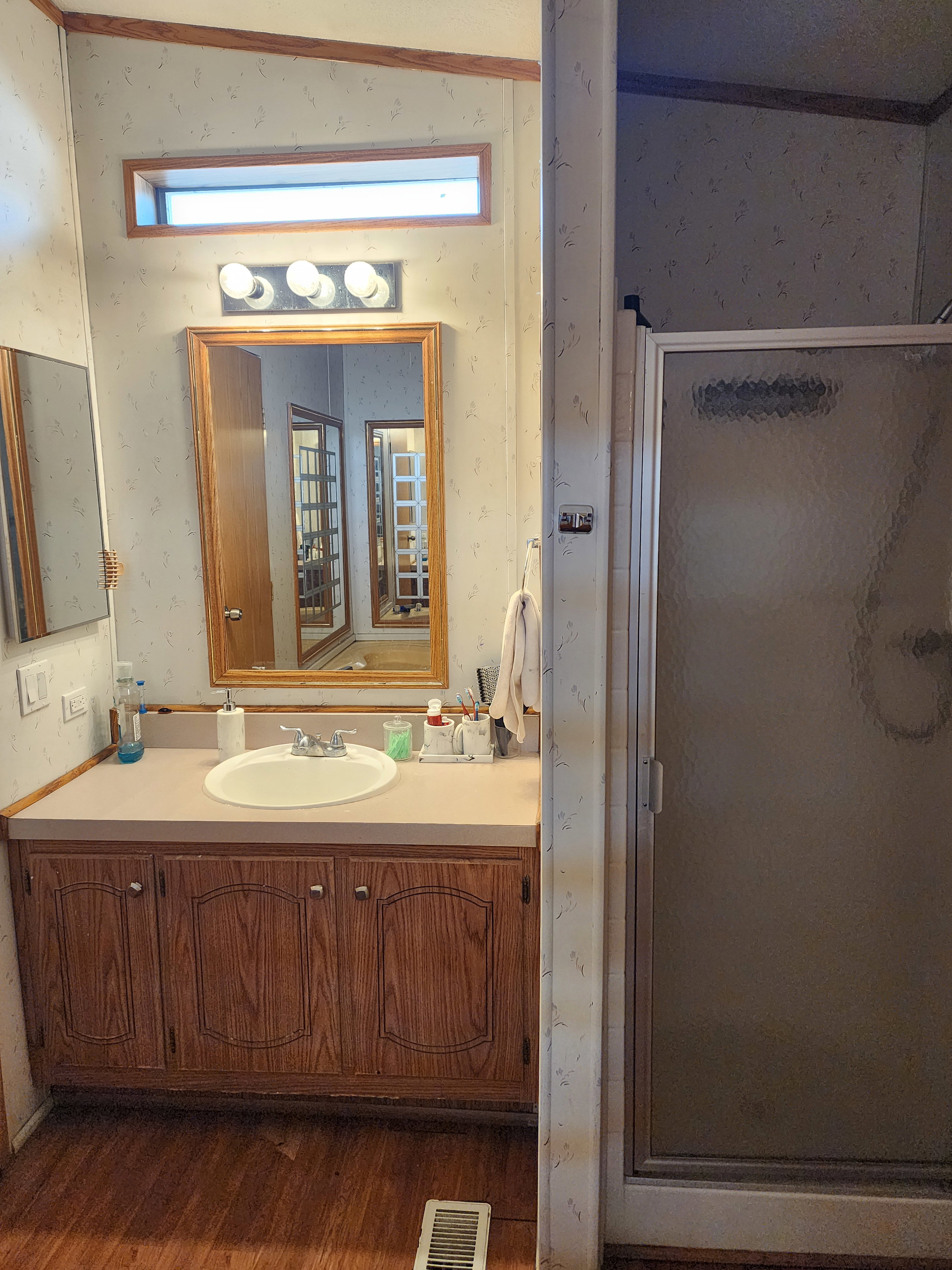 Bathroom featuring a wood-finish vanity with an integrated sink, a large framed mirror, overhead vanity lighting, a transom window, and wood-finish flooring
