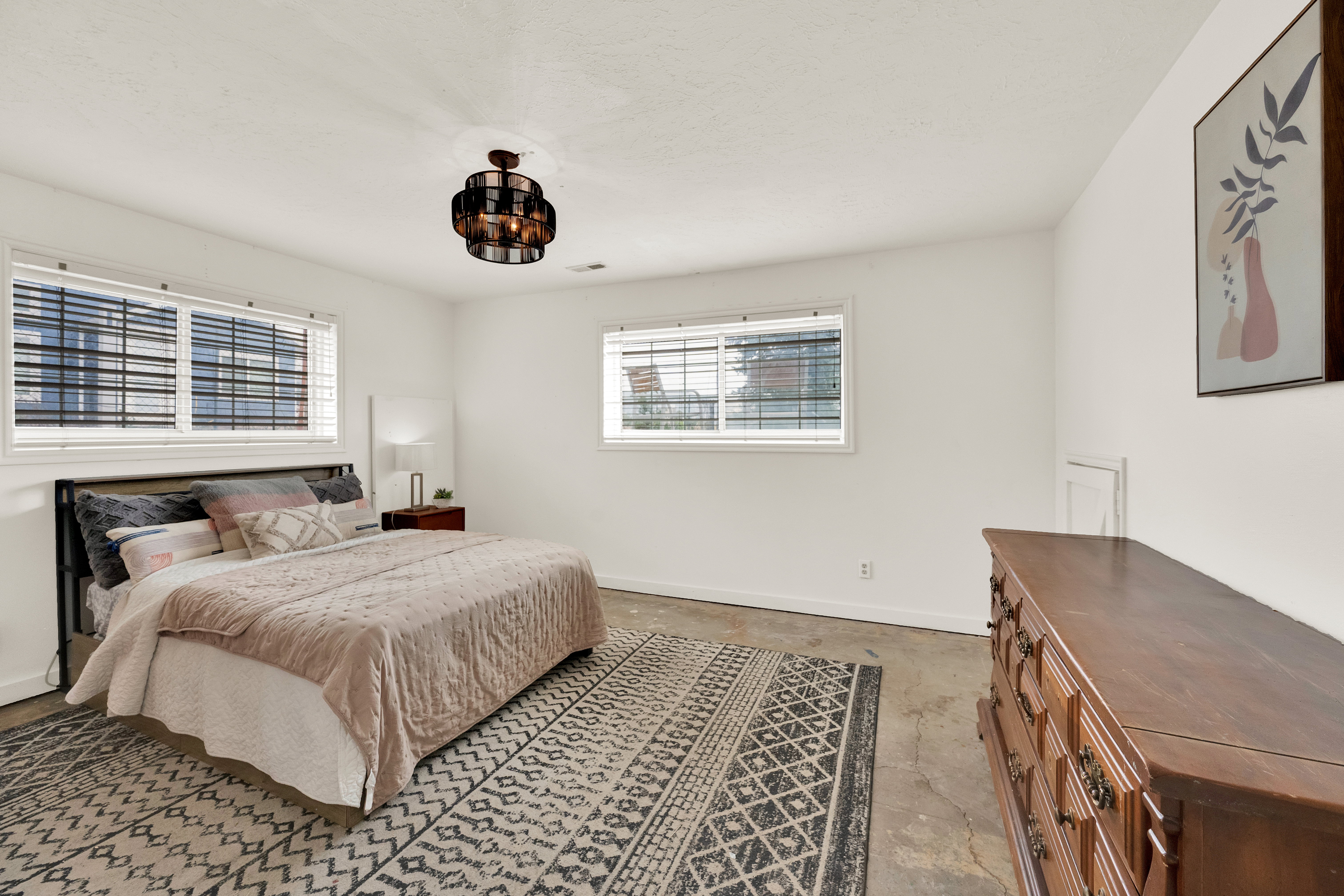 Bright bedroom featuring two windows with horizontal blinds, a ceiling-mounted fixture, and concrete flooring