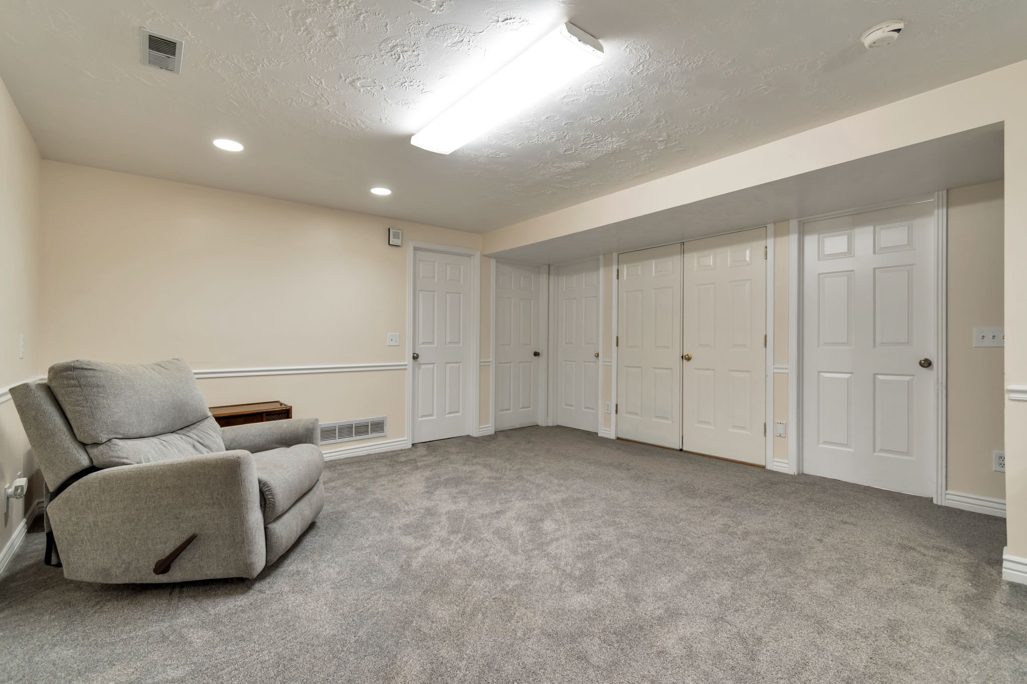 Spacious interior room featuring light-toned walls, grey carpeting, multiple white paneled doors, and recessed lighting with an additional fluorescent fixture