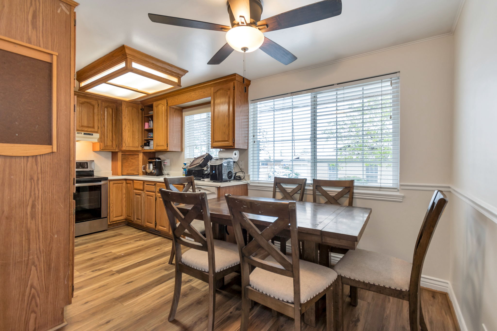 Kitchen and dining area featuring wood-finish flooring, wood cabinetry, stainless steel appliances, a ceiling fan with integrated lighting, and a large window with blinds