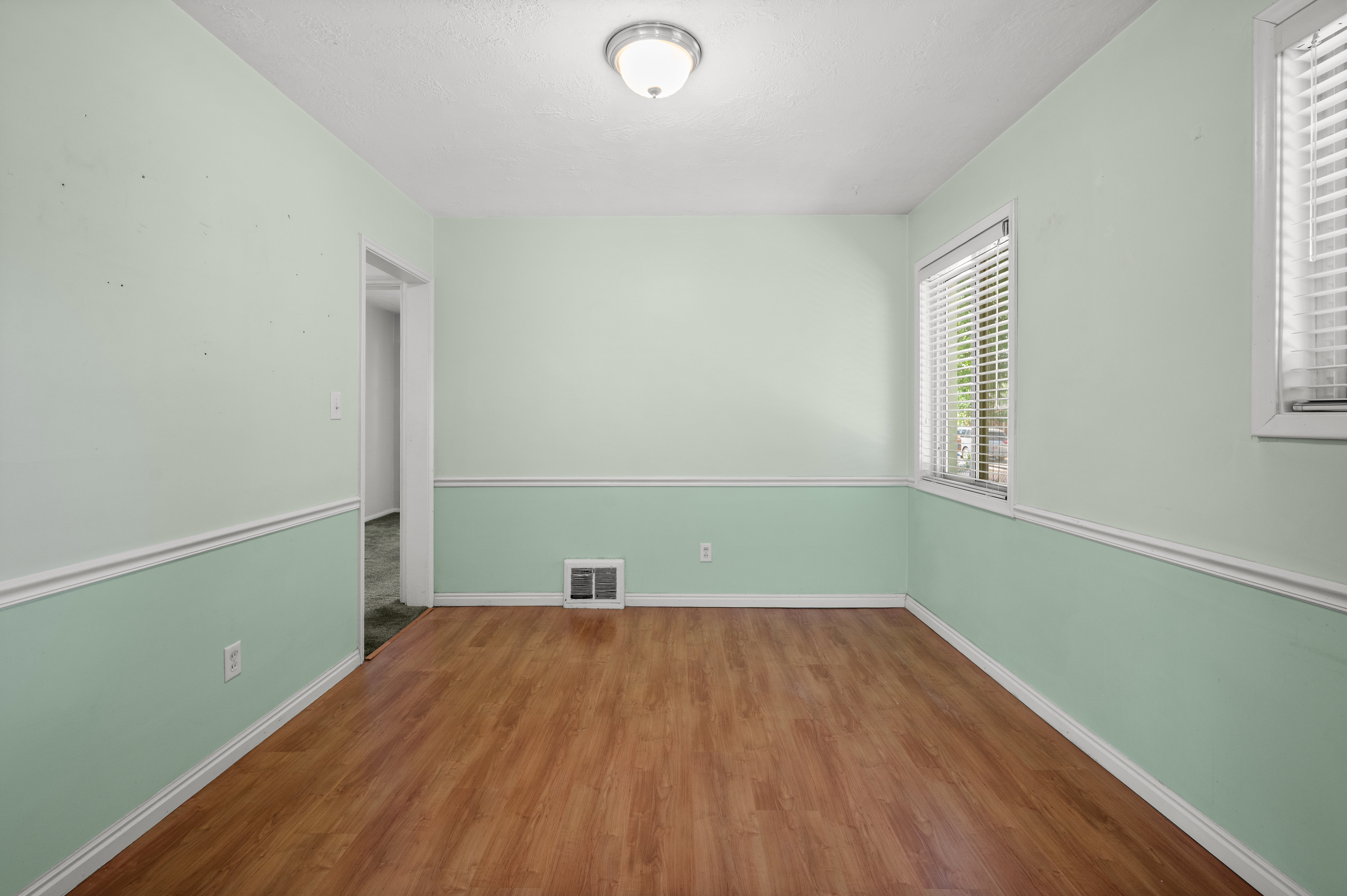 Spacious room featuring wood-finish flooring, two windows with blinds, a ceiling-mounted light fixture, and two-tone painted walls with chair rail molding