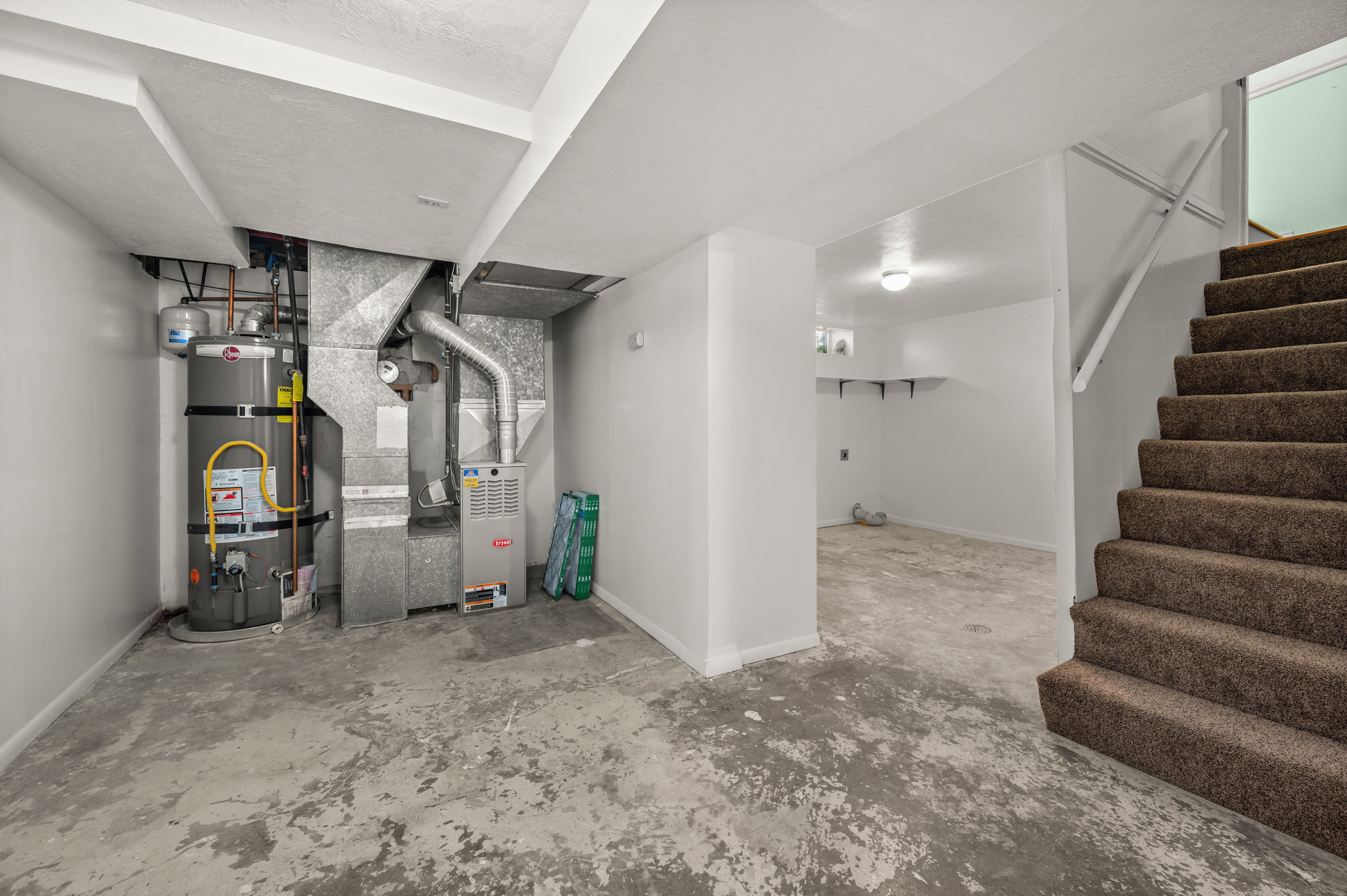 Spacious lower level featuring a utility area with water heater and furnace, exposed ductwork, carpeted staircase with white handrail, concrete flooring, and white painted walls