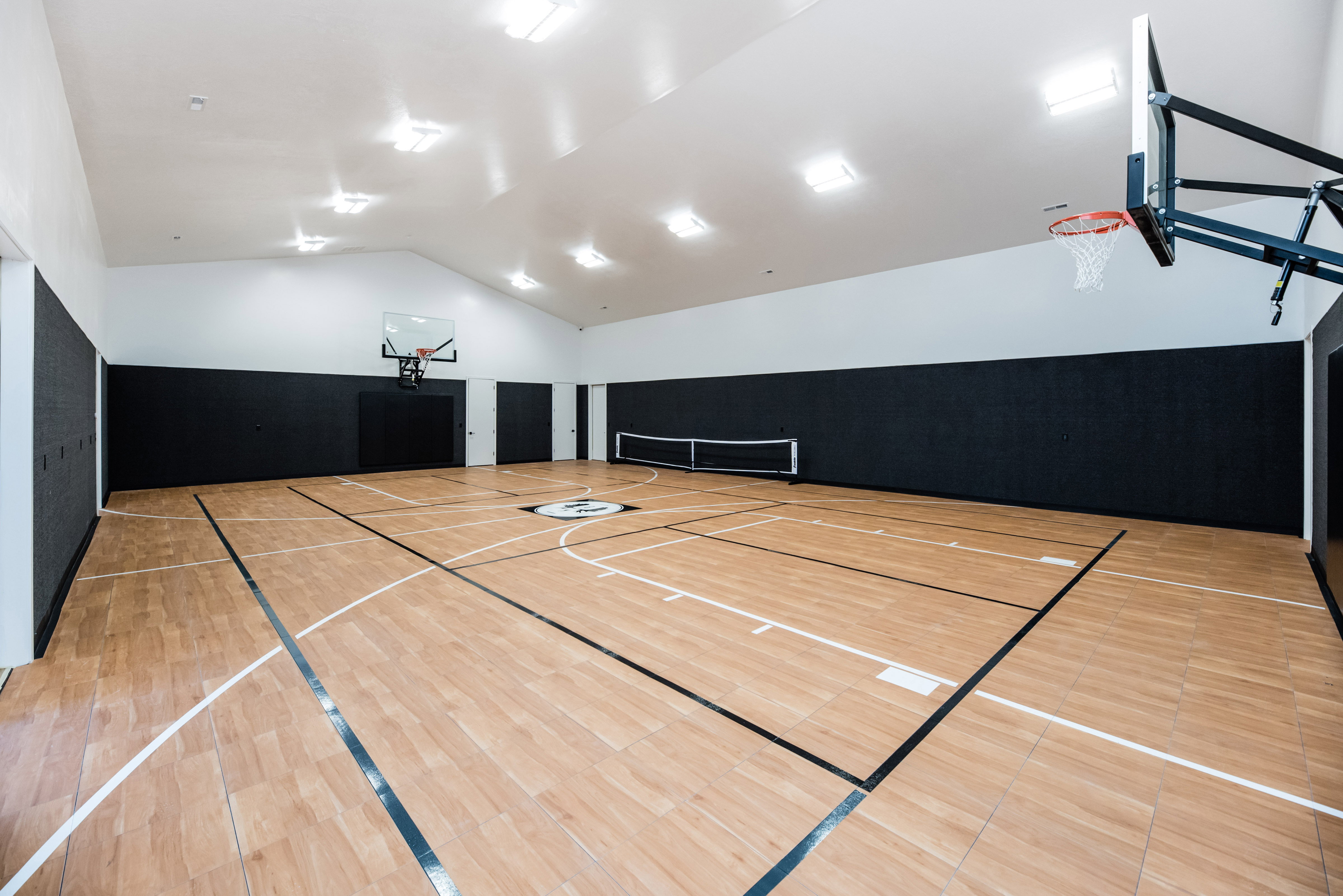 Spacious indoor athletic court featuring wood-finish flooring with court markings, dual basketball hoops, and a ping-pong net