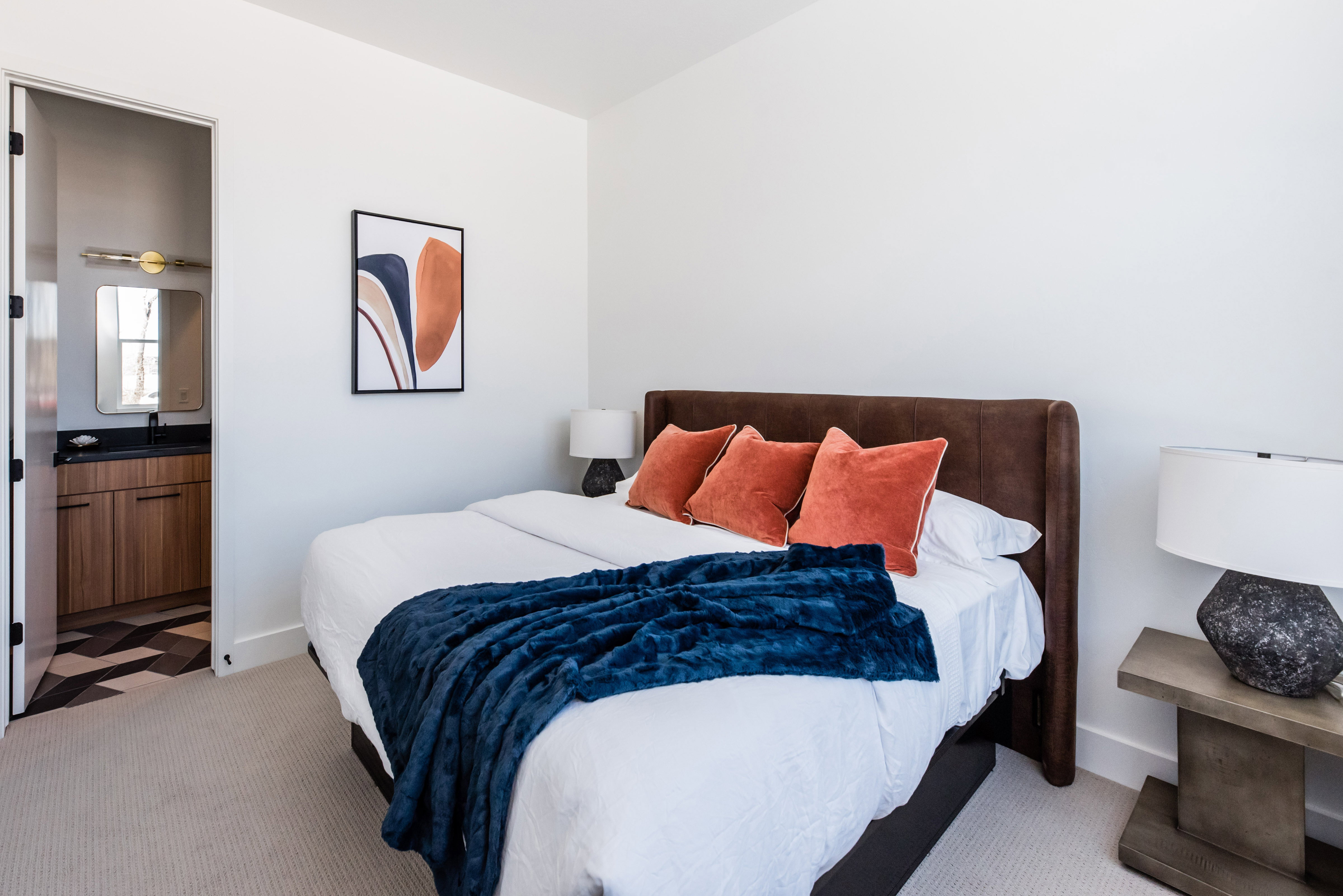 Carpeted bedroom with a built-in upholstered headboard, white walls, and a doorway revealing a bathroom with a wood-finish vanity and patterned floor tile