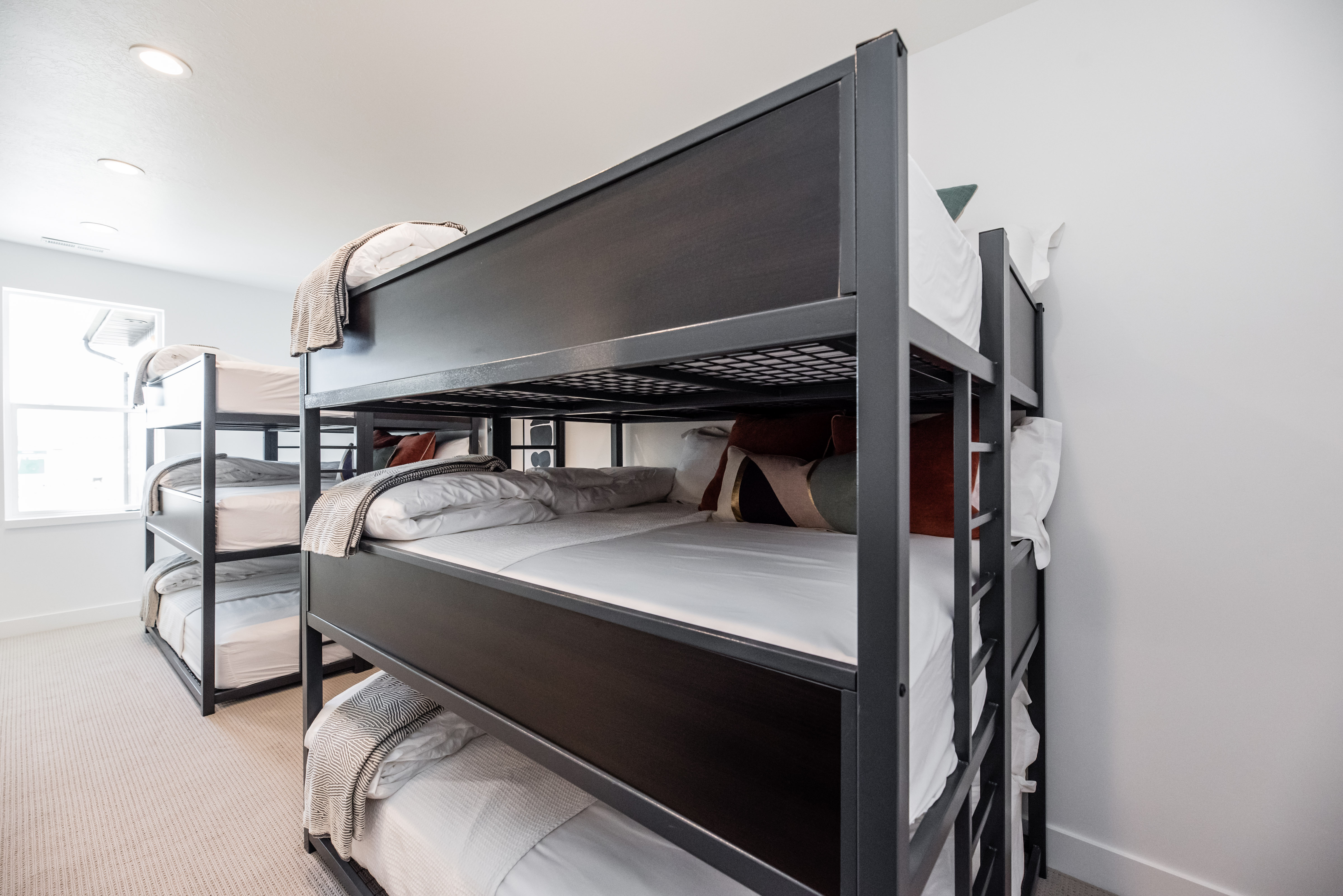 Spacious room featuring multiple bunk beds with metal frames, recessed ceiling lighting, a large window, light-toned carpeting, and crisp white walls