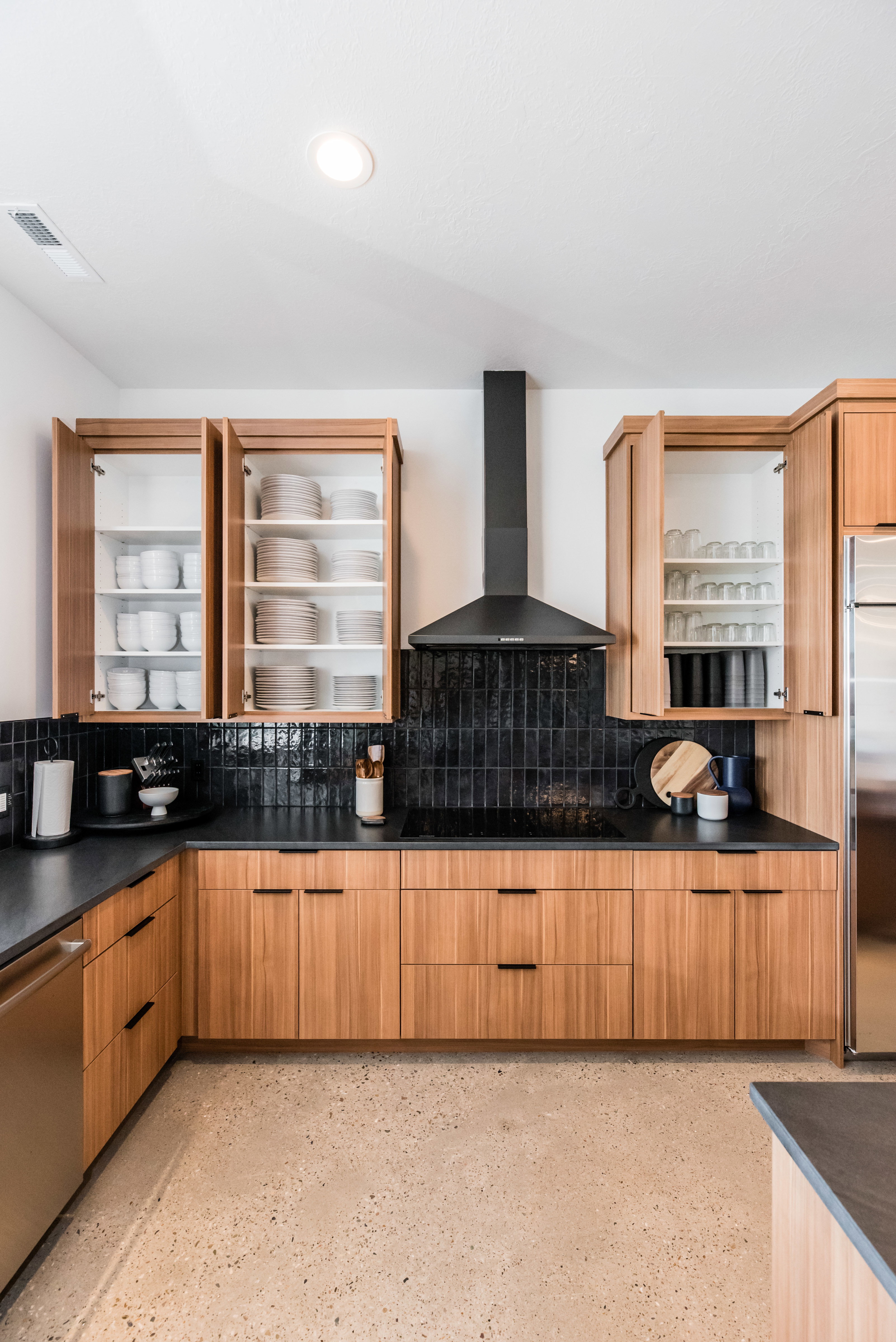 Contemporary kitchen featuring warm wood-finish cabinetry, dark countertops, and a full-height black tile backsplash