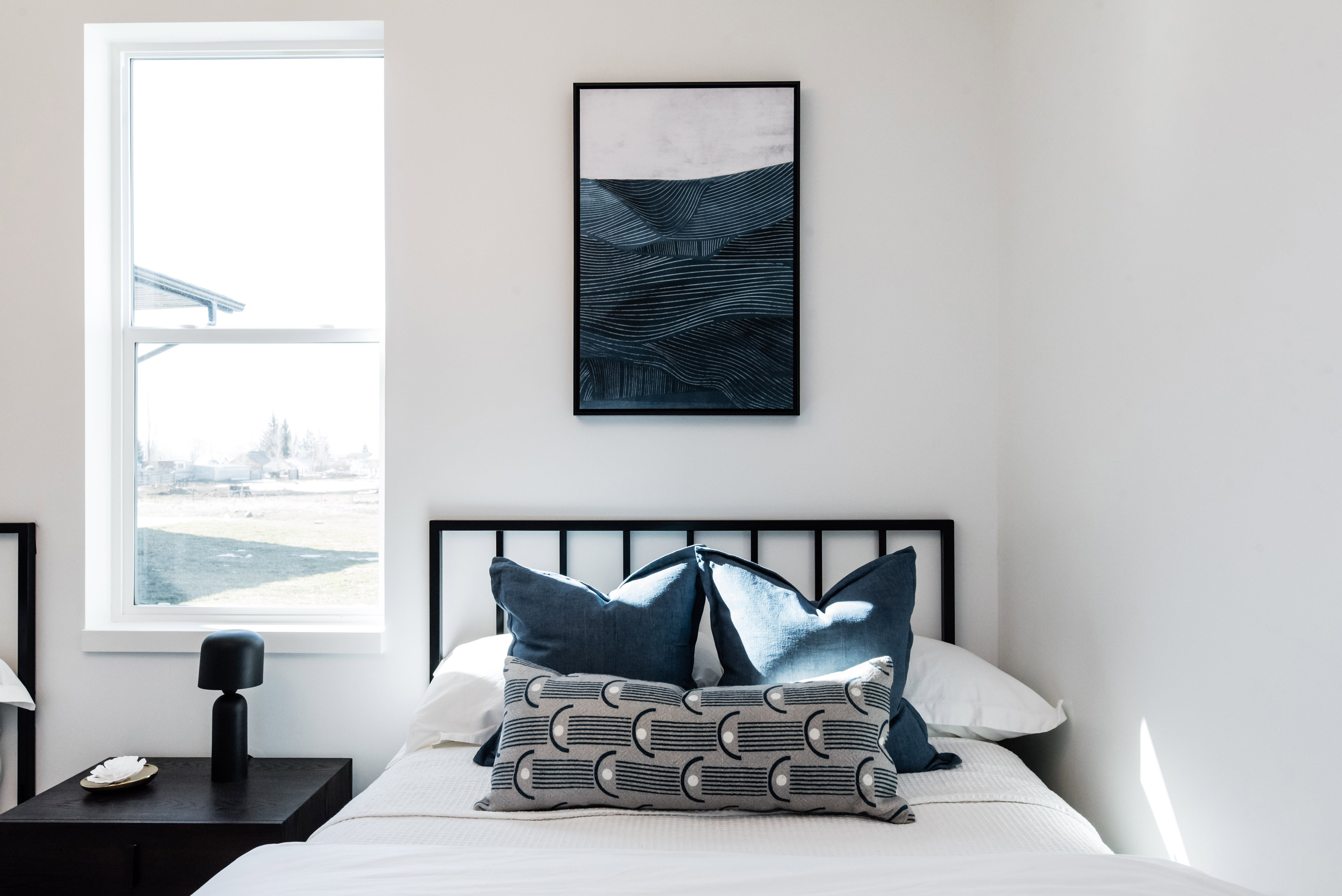White walls with a tall window, a black metal headboard, and a dark wood-finish nightstand