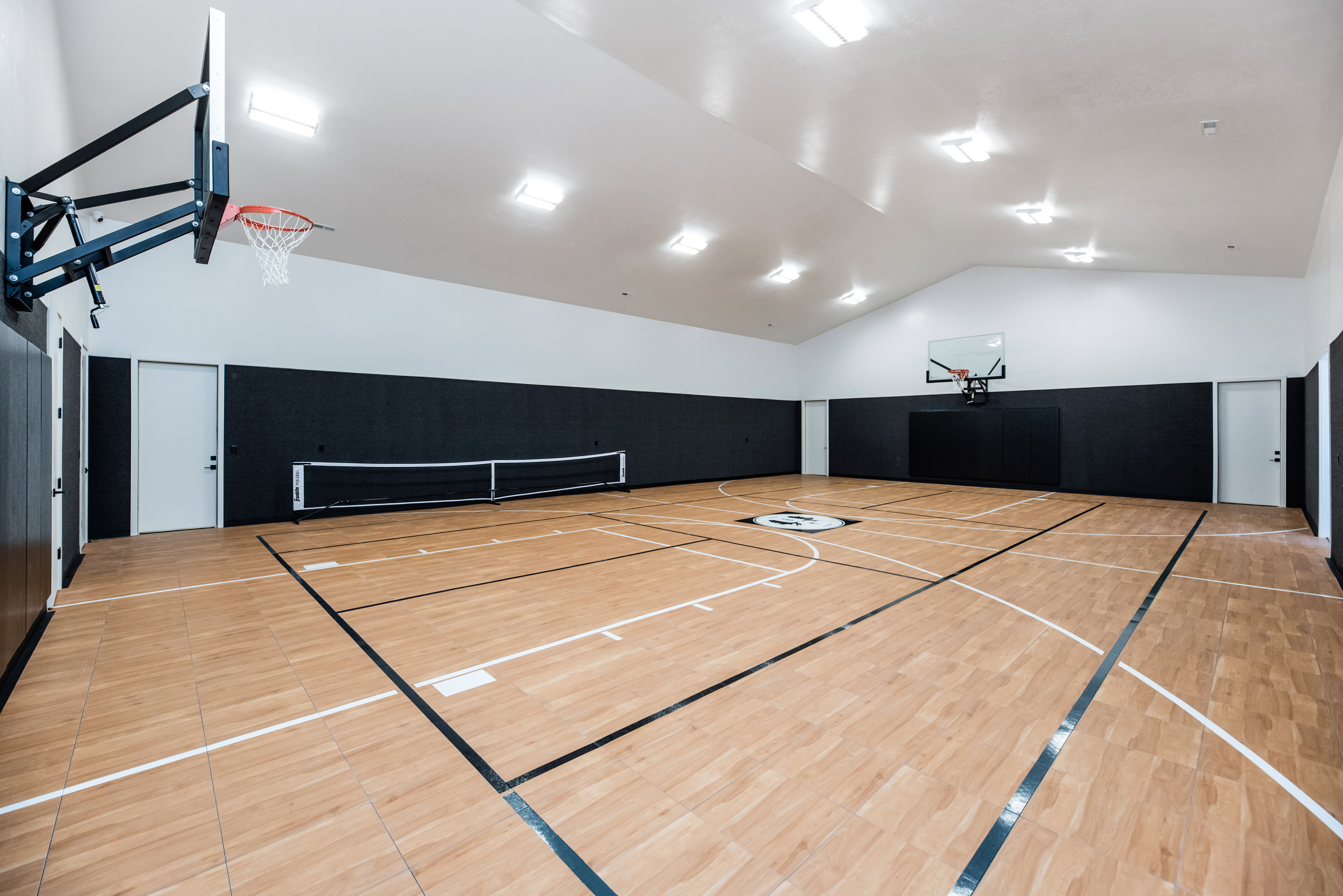 Indoor athletic court featuring wood-finish flooring with marked lines, two basketball hoops, and dark padded walls