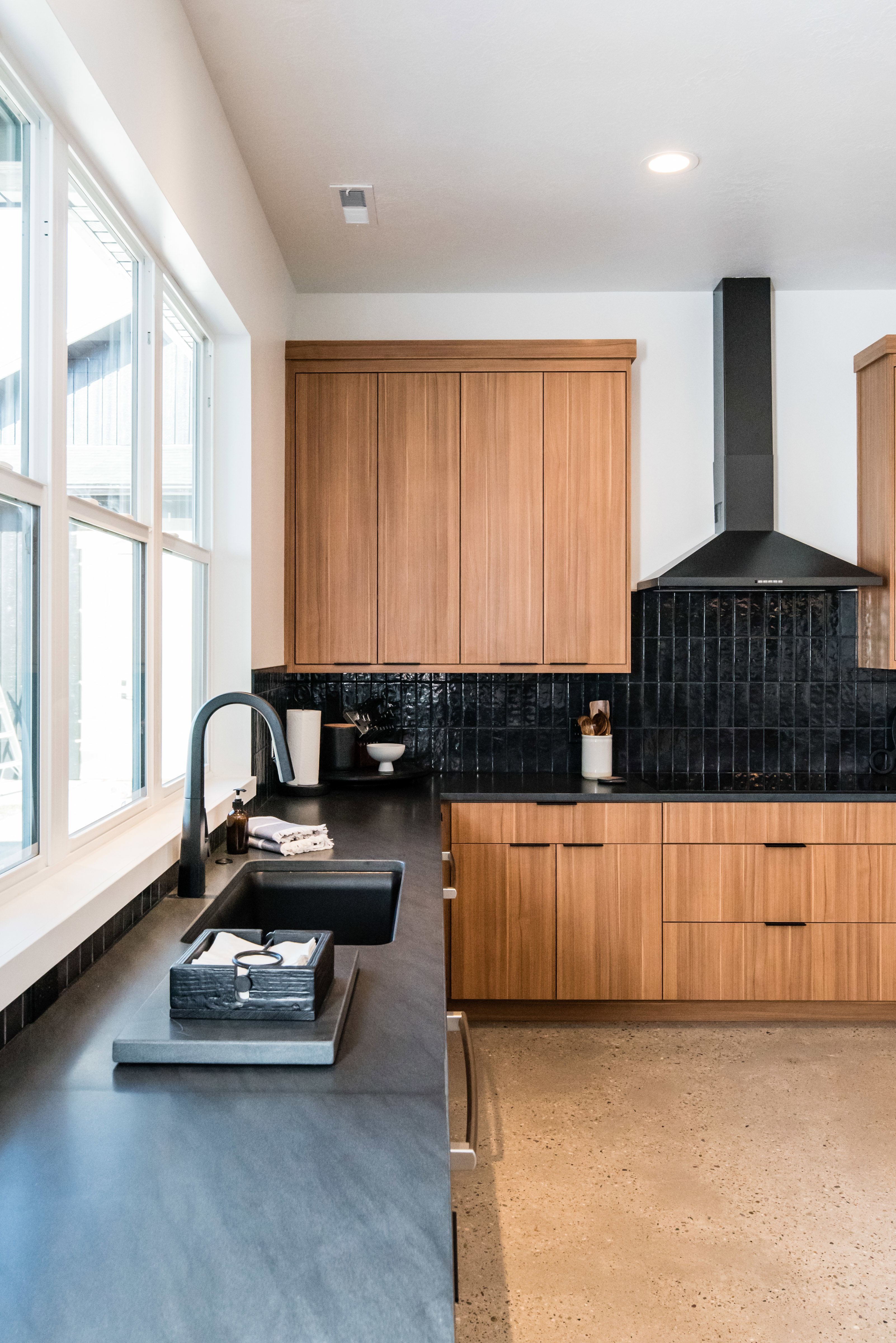 Modern kitchen featuring sleek wood-finish cabinetry, dark matte countertops, and a black tile backsplash