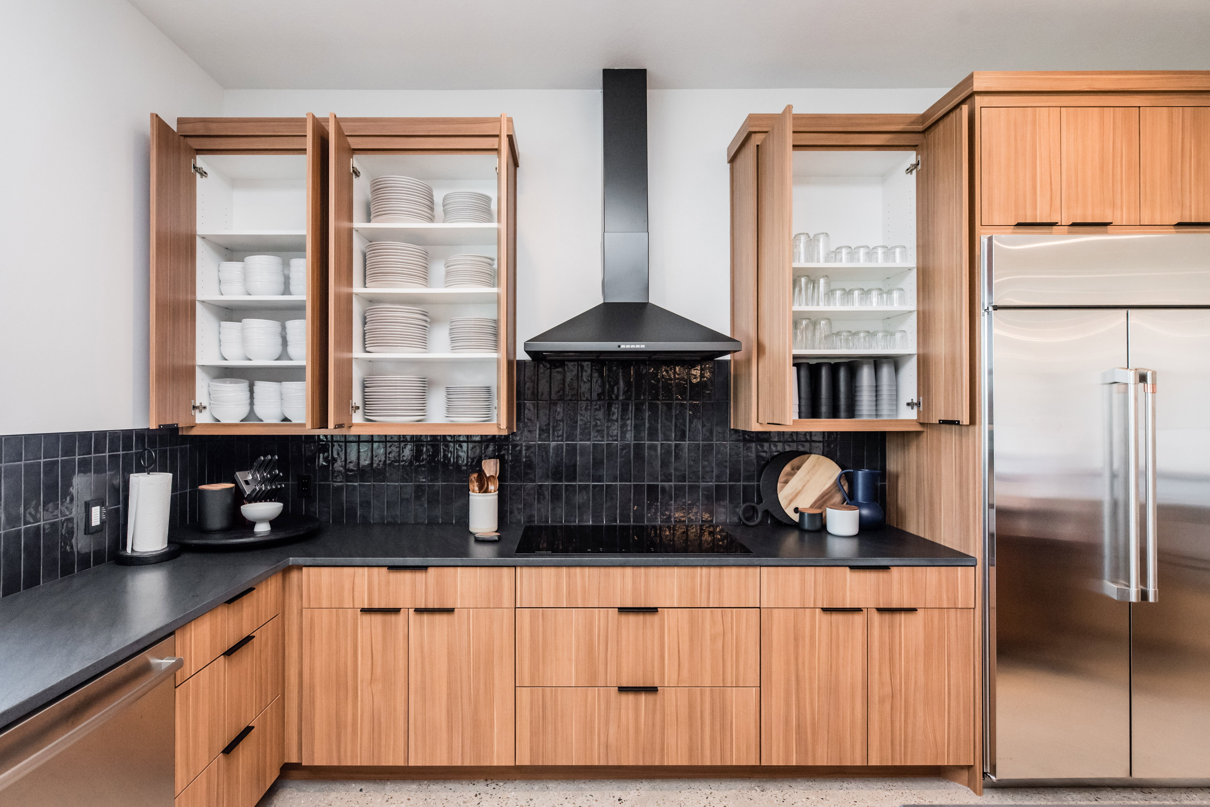 Modern kitchen featuring sleek wood cabinetry, black countertops, a black tile backsplash, and a stainless steel refrigerator