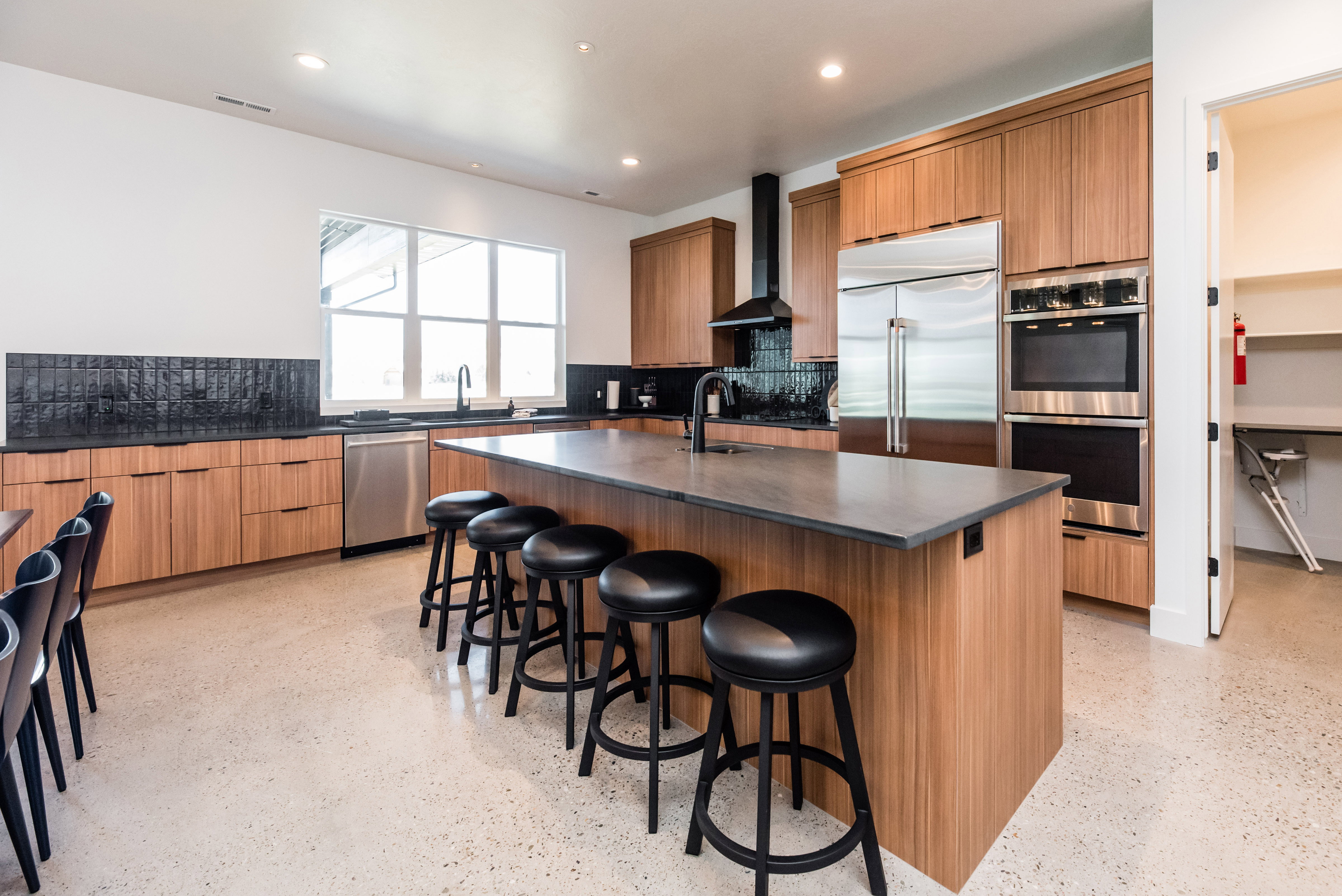 Modern kitchen featuring wood-finish cabinetry, a large center island with seating, stainless steel appliances including a double oven and refrigerator, a black tile backsplash, and a spacious walk-in pantry