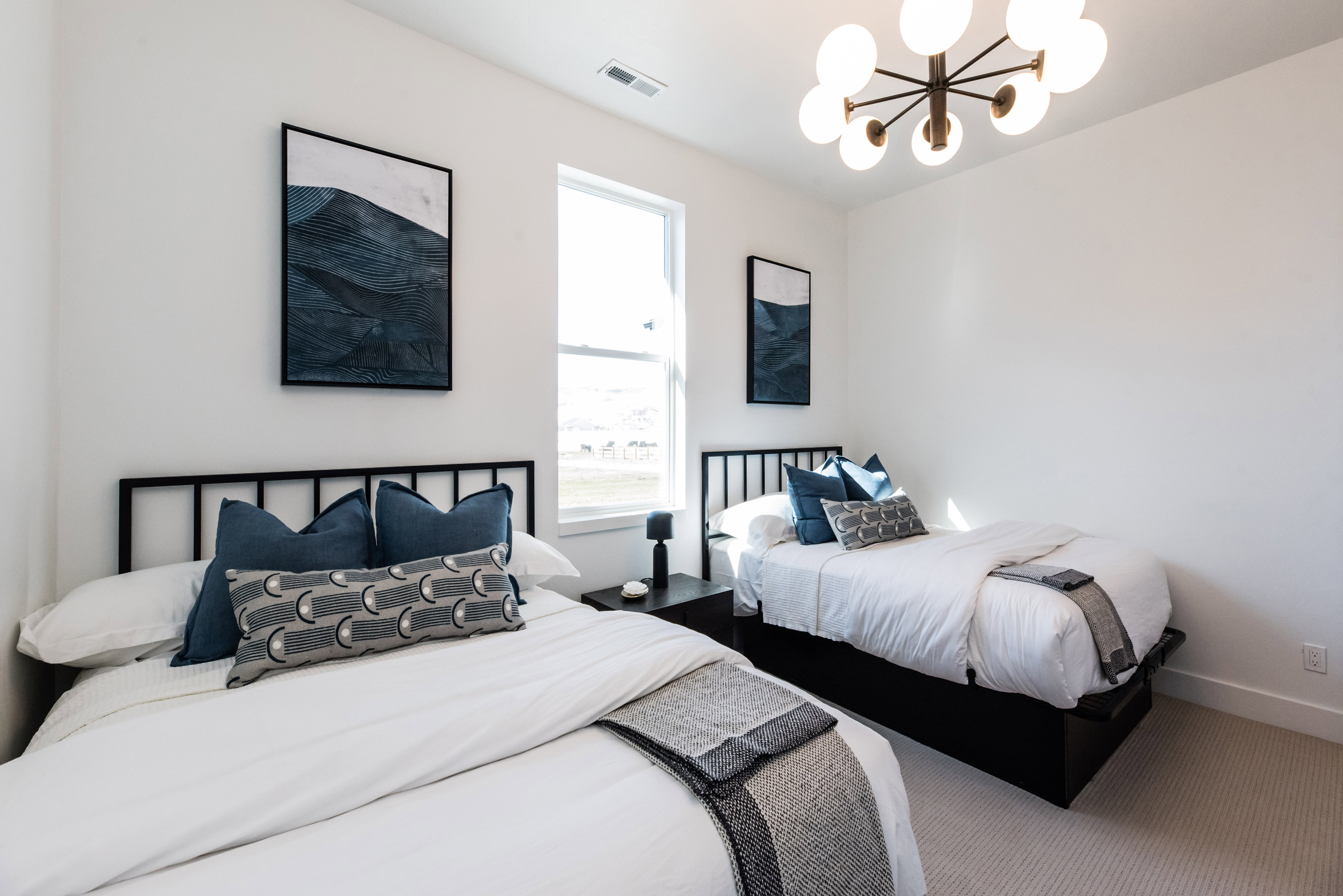 Bright room featuring two beds with black metal headboards, a large window, a modern chandelier, and light-toned carpet flooring
