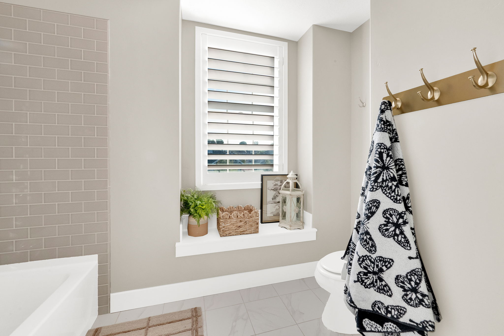 Recessed window with plantation shutters, light gray subway tile shower surround, white baseboards, and wall-mounted brass coat hooks