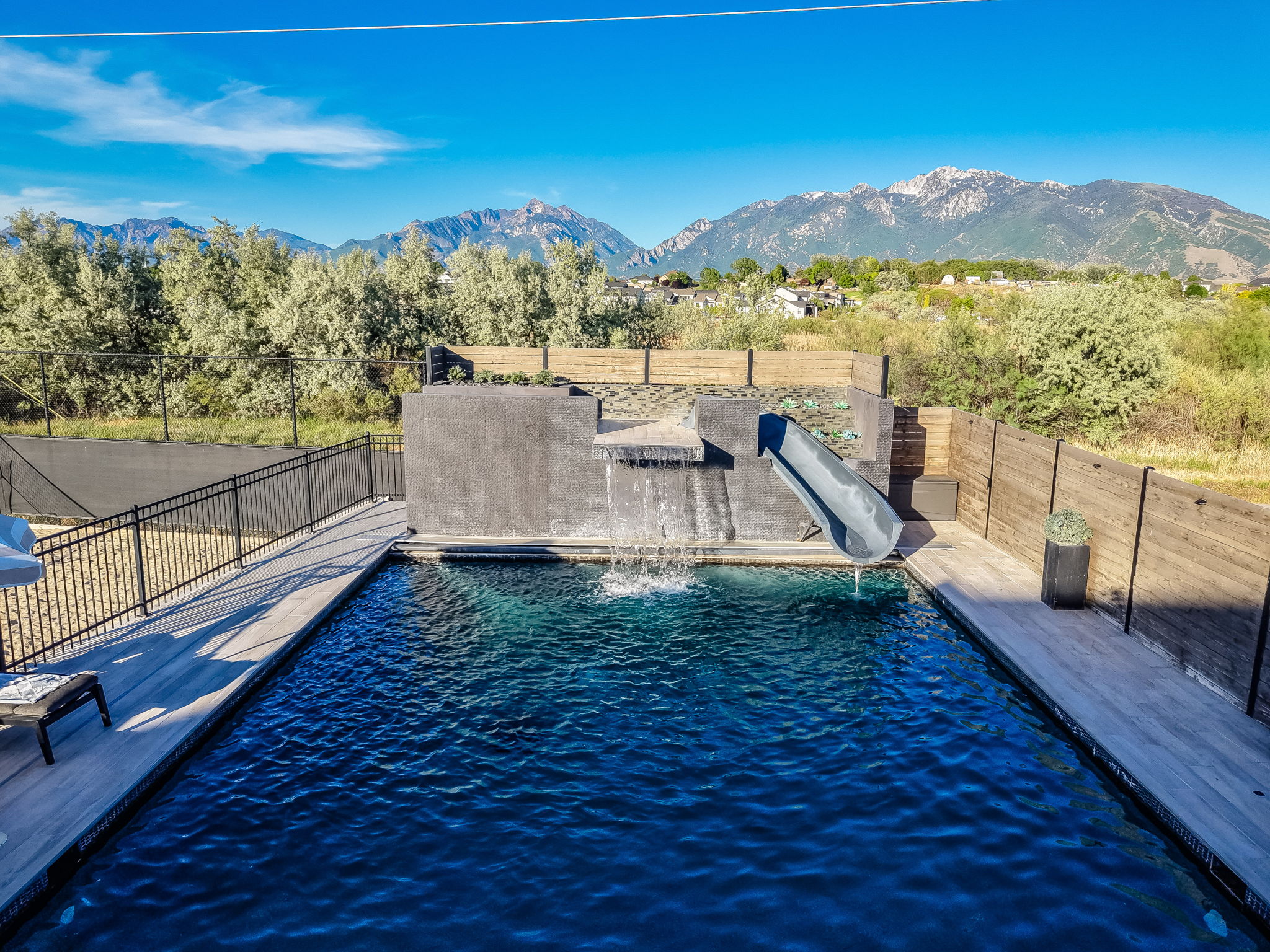 Outdoor swimming pool with a dark finish, integrated slide, waterfall feature, and surrounding wood-finish decking