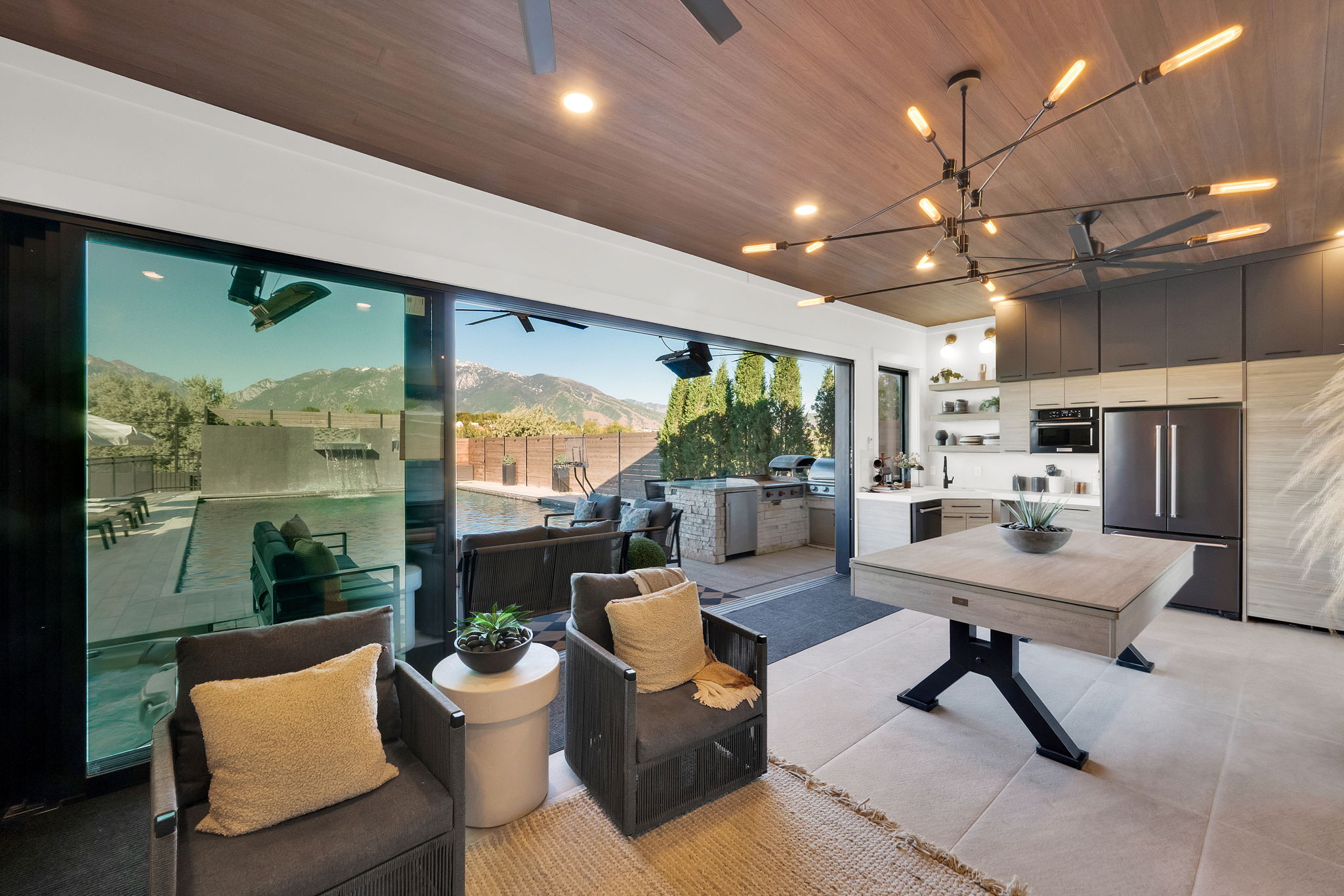 Expansive indoor-outdoor living space featuring floor-to-ceiling sliding glass doors, a wood-finish ceiling, and a modern linear chandelier