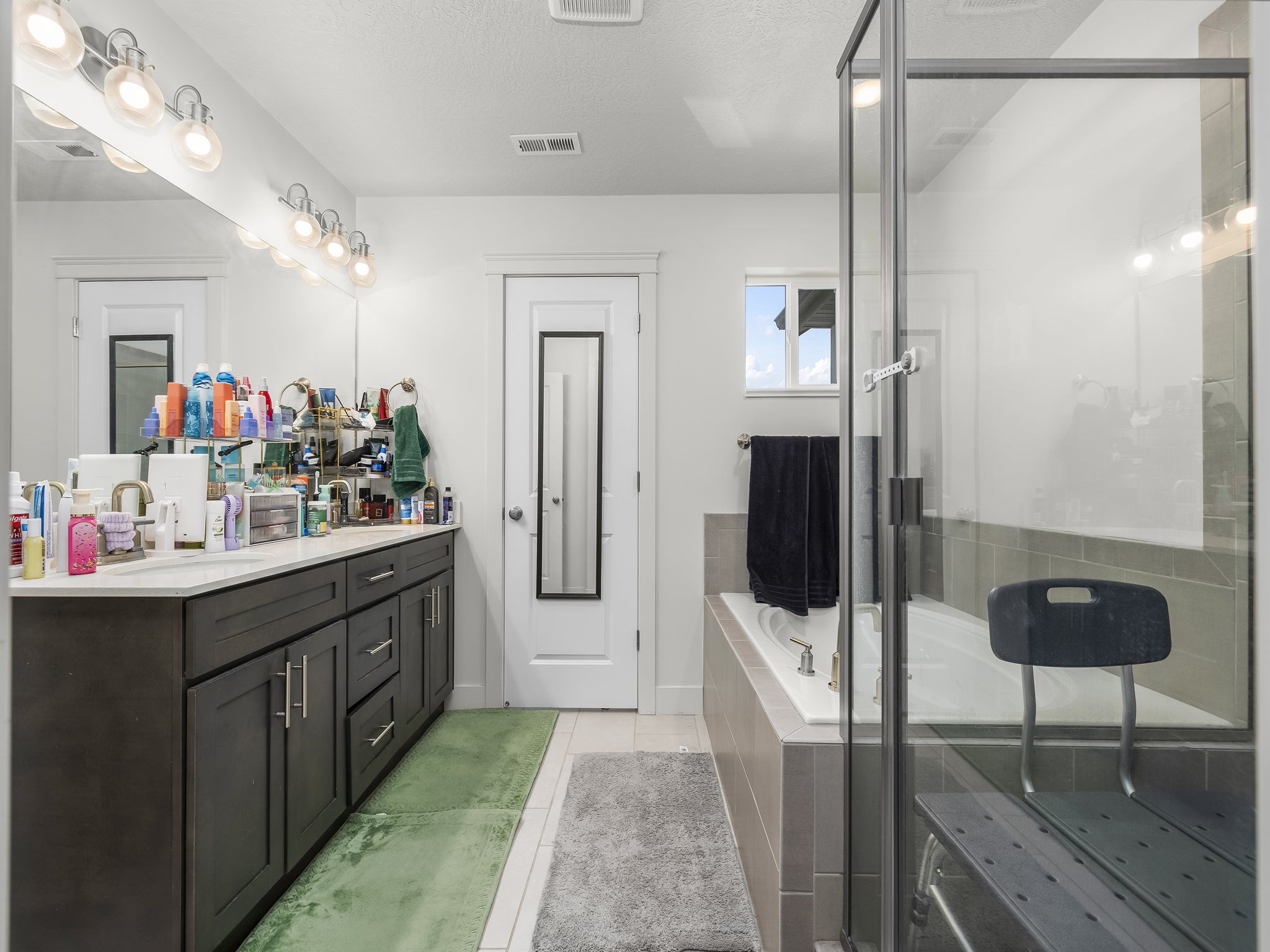 Bathroom featuring a dual vanity with dark wood-finish cabinetry, a large frameless mirror, a built-in soaking tub with tiled surround, and a glass-enclosed shower
