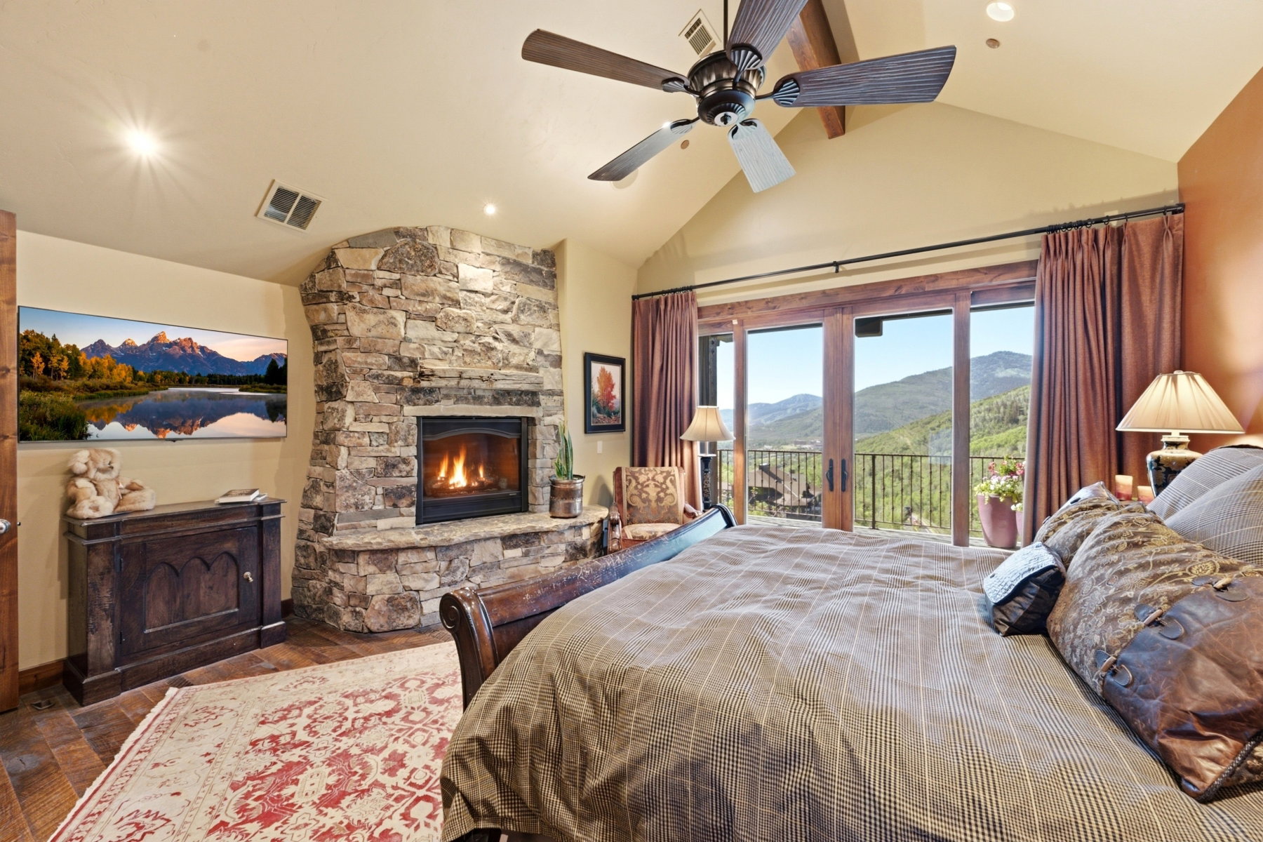 Stone-clad corner fireplace with hearth, wood-finish flooring, vaulted ceiling with ceiling fan, and French doors opening to mountain scenery