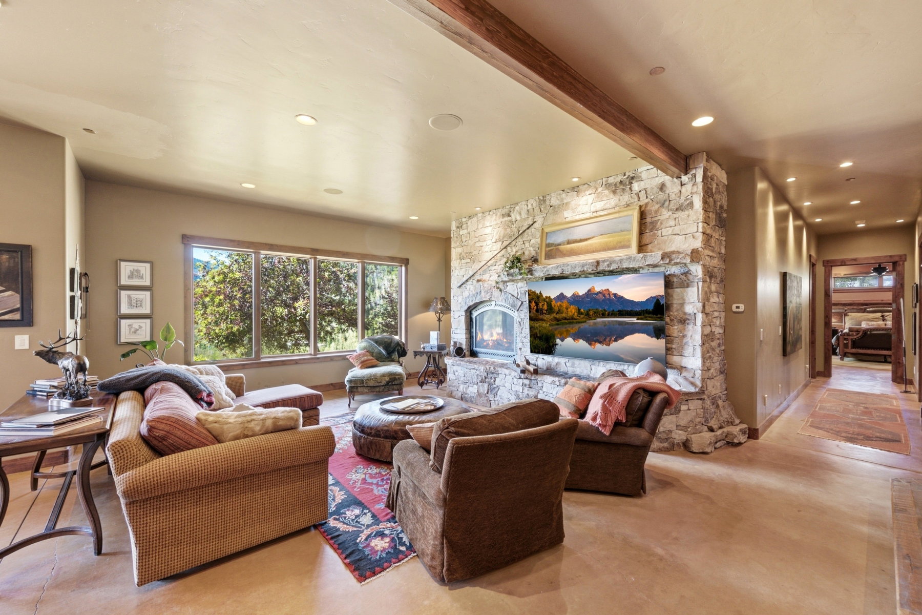 Spacious living area featuring a stone-clad fireplace and hearth, a large picture window, exposed wood beams, and recessed lighting
